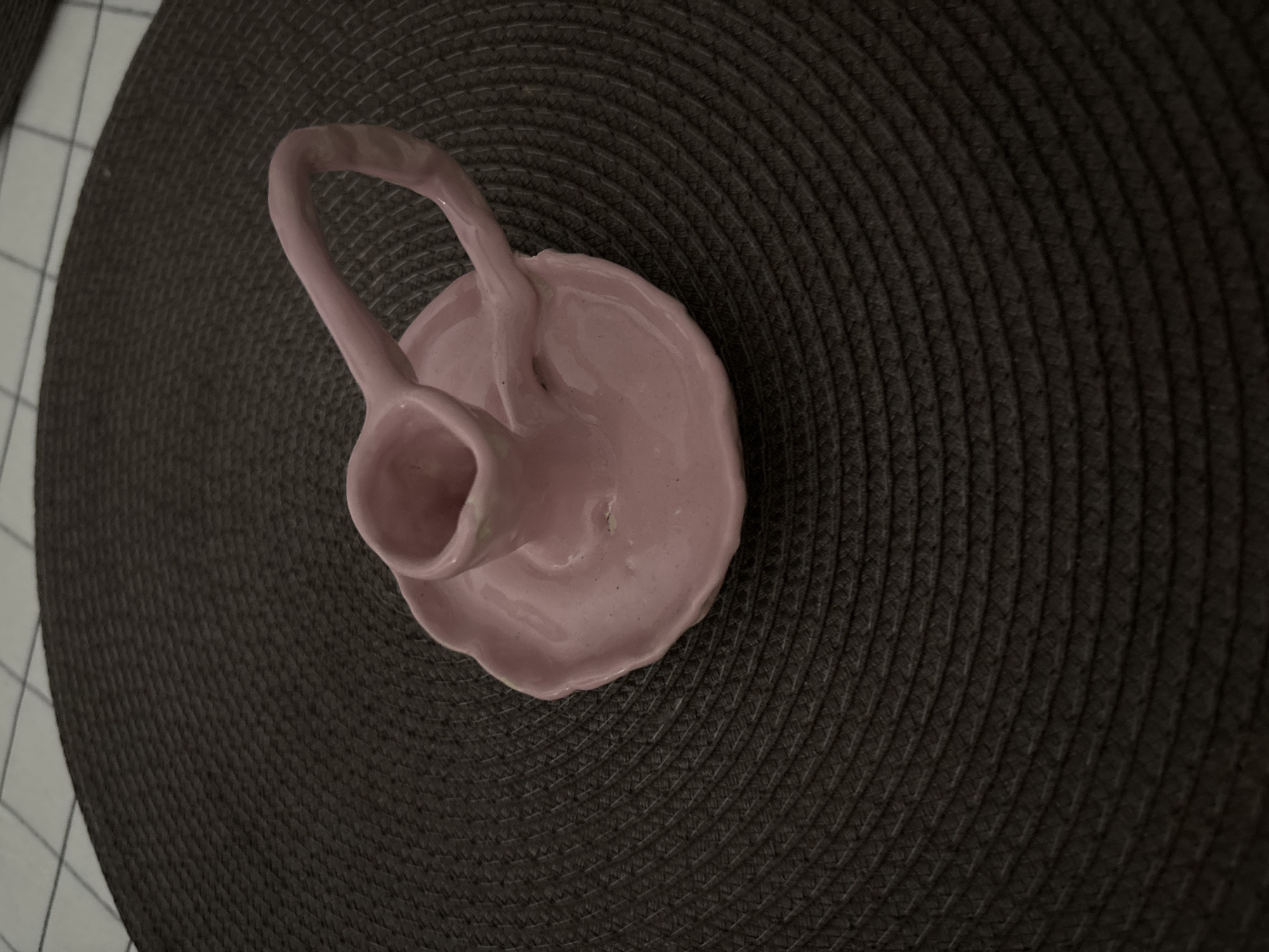 The image shows a pink ceramic object with a handle, possibly a decorative vase or candle holder, placed on a dark brown woven placemat. The placemat sits on a surface with a white and gray checkered pattern, which suggests a kitchen or dining area. The lighting is subdued. No specific location in Saint Petersburg, Russia, can be determined from the image. There are no people visible in the scene.