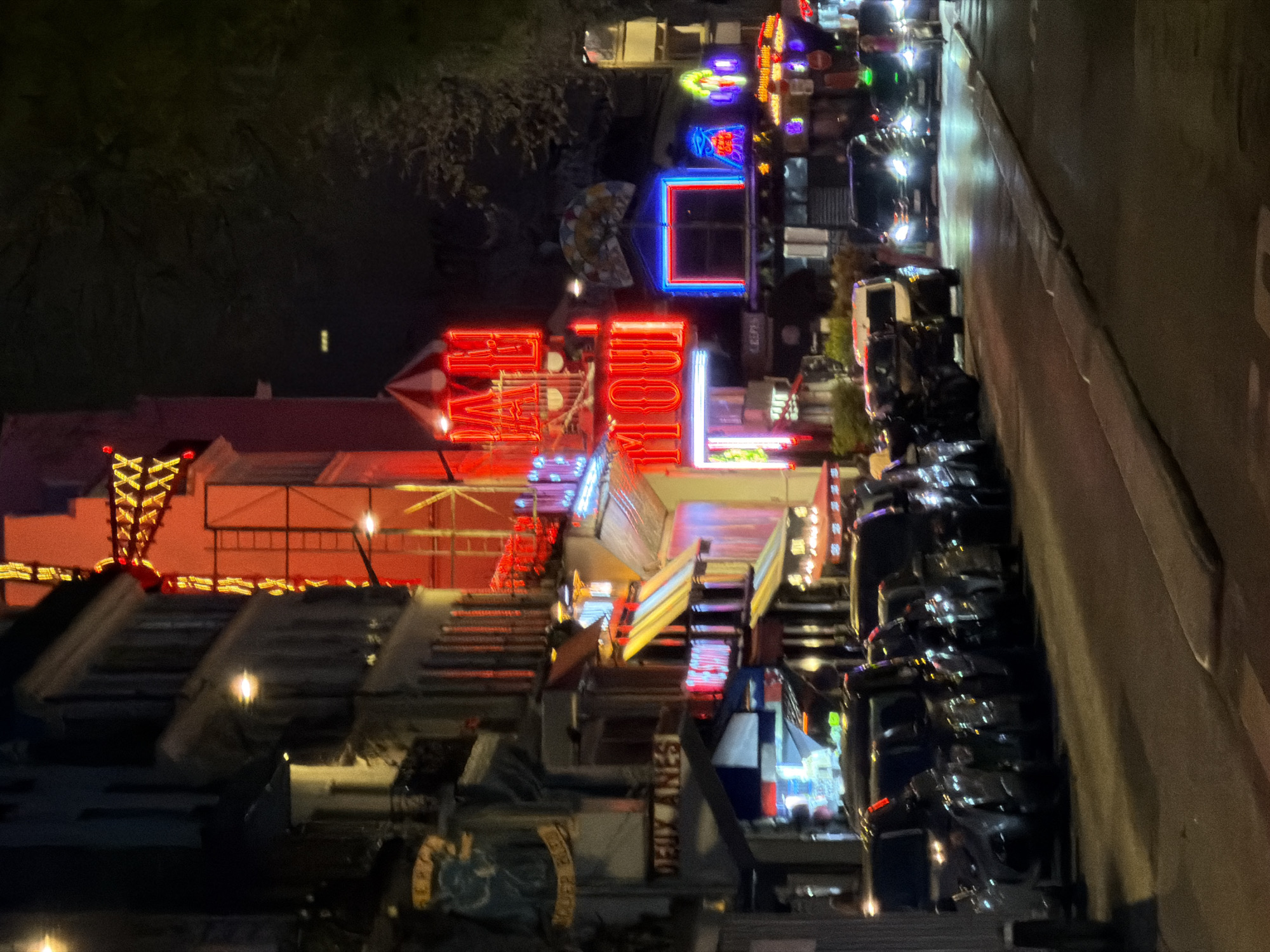 The image depicts a street scene in Paris, France at night. The focus is on the vibrant and colorful displays of the Moulin Rouge. Other buildings, including a restaurant named “Deux Anes,” are located in the foreground. Cars are parked along the right side of the street. Tree limbs overhang the left side of the scene.