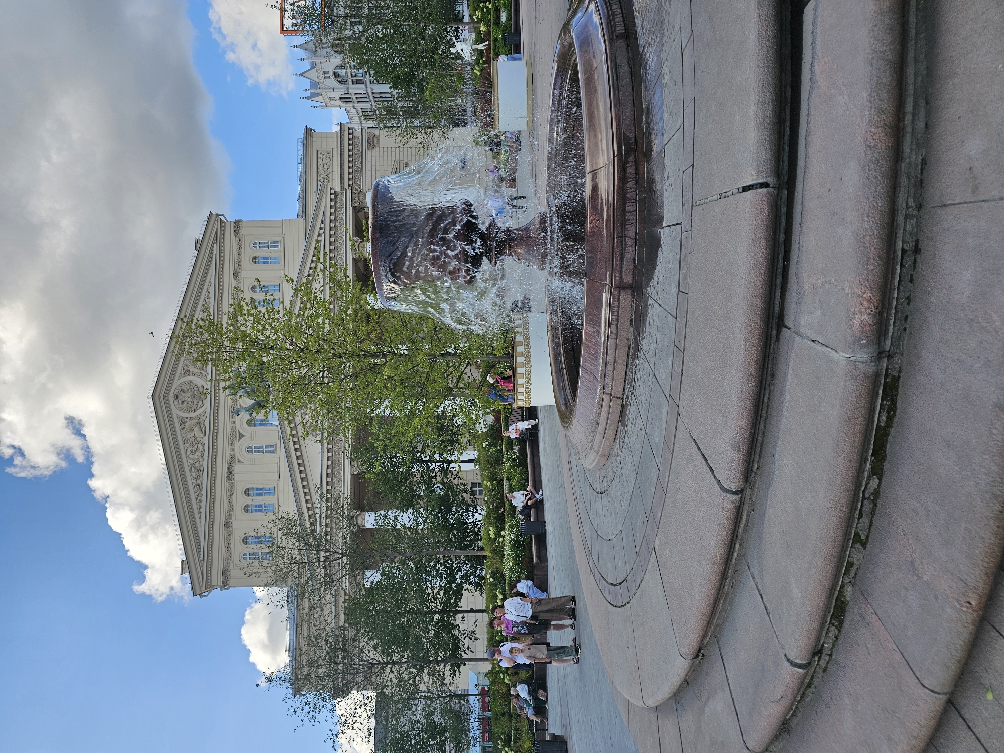 A wide shot captures a fountain in the foreground, with water spraying upwards from a large, ornate basin. Behind the fountain, a grand, classical building with a pedimented facade stands under a partly cloudy blue sky. Trees line the walkway in front of the building, and several people are seated on benches or standing in small groups. The fountain is made of dark stone and is surrounded by a paved area with curved edges. The building has a light-colored facade with numerous windows and decorative elements. The overall scene appears to be a public square or park in an urban setting.