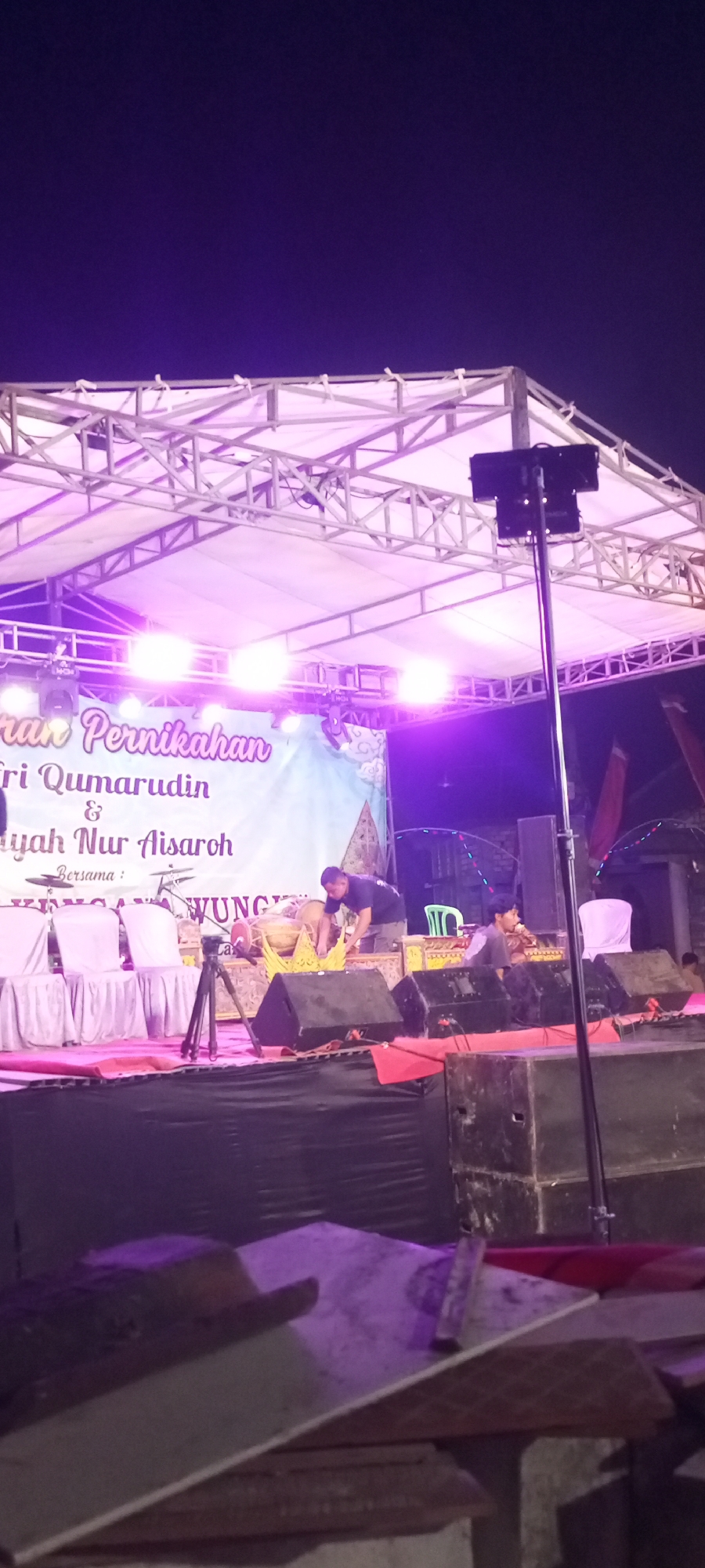 The image depicts an outdoor stage at night, illuminated by purple and pink lights, possibly for a wedding reception in Paji, Indonesia. The stage features a white tent-like covering supported by a metal truss structure. Several spotlights are visible on the truss. On the stage, a banner with Indonesian text, likely announcing a wedding, is displayed. Two men are present; one is adjusting musical instruments, including what appear to be traditional drums, while the other is seated and observing. A tripod and speakers are also on the stage. In the foreground, wooden planks and a tiled surface are visible, suggesting construction or debris. The background shows a dark sky with some distant lights and structures.