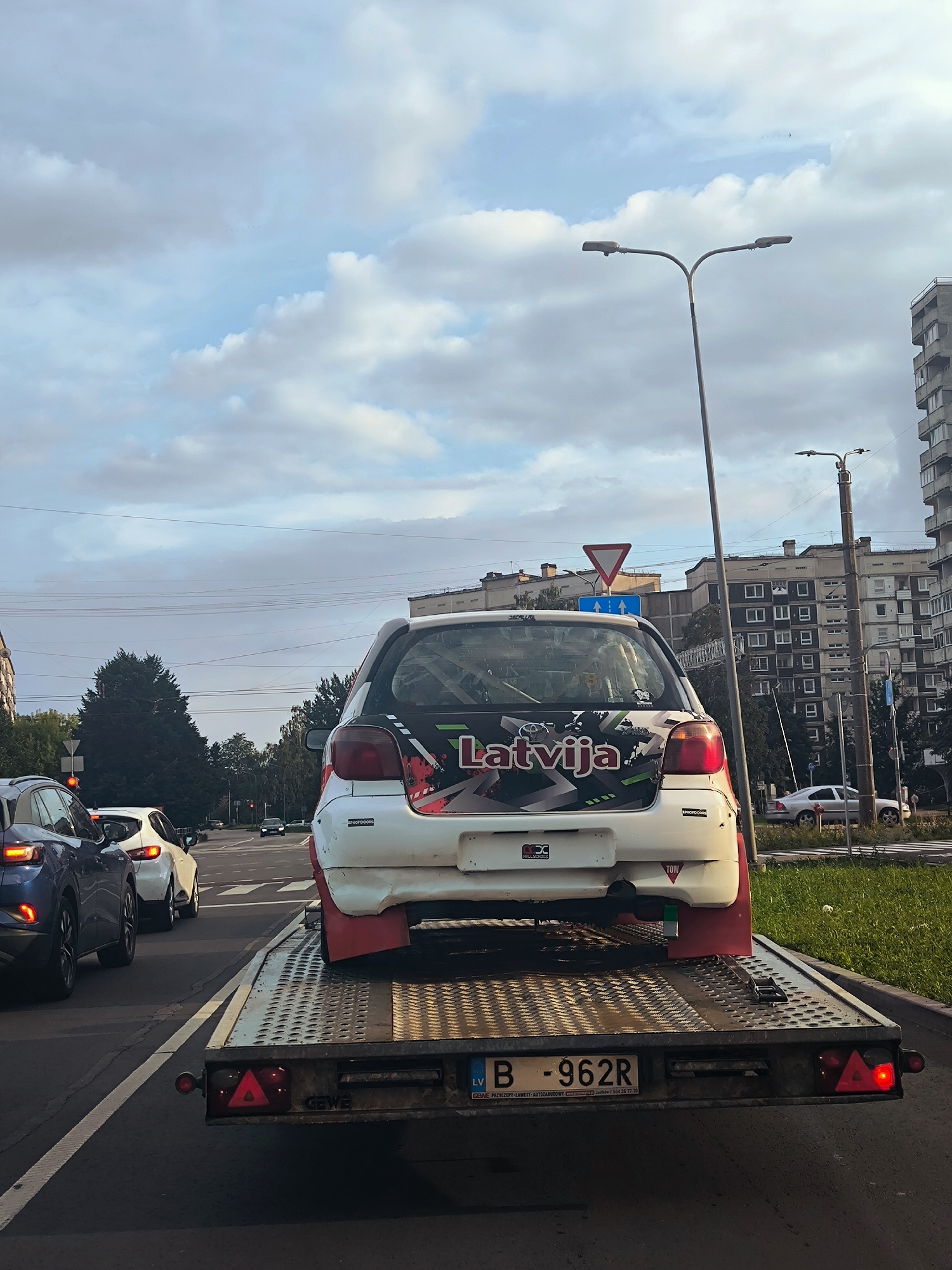In Riga, Latvia, on a street lined with apartment buildings and trees, a white car, likely a Toyota Yaris, is being transported on a flatbed trailer. The car is decorated with a large "Latvija" decal and what appears to be racing graphics. It has prominent red mudguards. The trailer itself has visible brake lights and a license plate reading "B - 962R". Other vehicles, including a dark blue SUV and a white sedan, are visible in traffic ahead. The sky is partly cloudy, with a street light and overhead wires present in the scene.