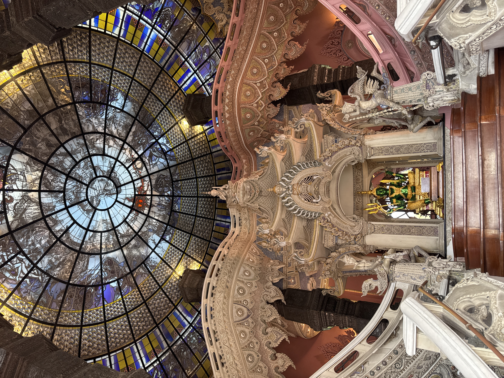The image depicts the interior of a temple in Samrong Nuea Subdistrict Municipality, Thailand. A large, circular stained-glass dome dominates the left side of the image, its panes filled with maps and vibrant colors. To the right of the dome is a highly ornate, multi-tiered structure with intricate carvings and statues, featuring a green Buddha statue on the upper level. The color palette is dominated by white, beige, and subtle pastel tones, accented by the saturated colors of the stained glass and the golden hue of the Buddha.