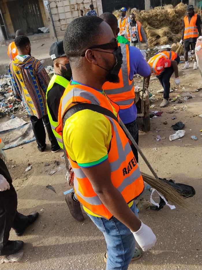 The image depicts a community clean-up operation in Fagge C, Nigeria, on an unpaved street littered with debris. The primary subject, a male, is positioned in the foreground, facing right in profile. He wears a bright yellow short-sleeved shirt with a multi-colored trim on the left sleeve, blue jeans, brown shoes, and white disposable gloves. Over his shirt, he wears an orange high-visibility safety vest with grey reflective stripes and the partial black text "RATEL" visible on his right chest area. He has dark-rimmed sunglasses, a black face mask covering his mouth and chin, and a neatly trimmed beard. He holds a traditional broom made of dried plant fibers in his right hand, angled towards the ground.
Behind the main subject, several other individuals, predominantly male, are engaged in similar activities or observing. One male directly behind the main subject wears a purple t-shirt and an orange safety vest. A wheelbarrow or handcart is partially visible between them. To the left, another male wears a yellow and blue patterned long-sleeved shirt and black trousers, walking away from the camera. Further into the scene, multiple men in orange safety vests are visible. One man in the mid-ground right is bent over, actively clearing debris from the ground next to a pile of dried plant material. The street surface is a mix of dirt and scattered trash, including plastic bags, paper, and various discarded items. Faded, utilitarian buildings with metal doors line the background, indicating an urban setting.