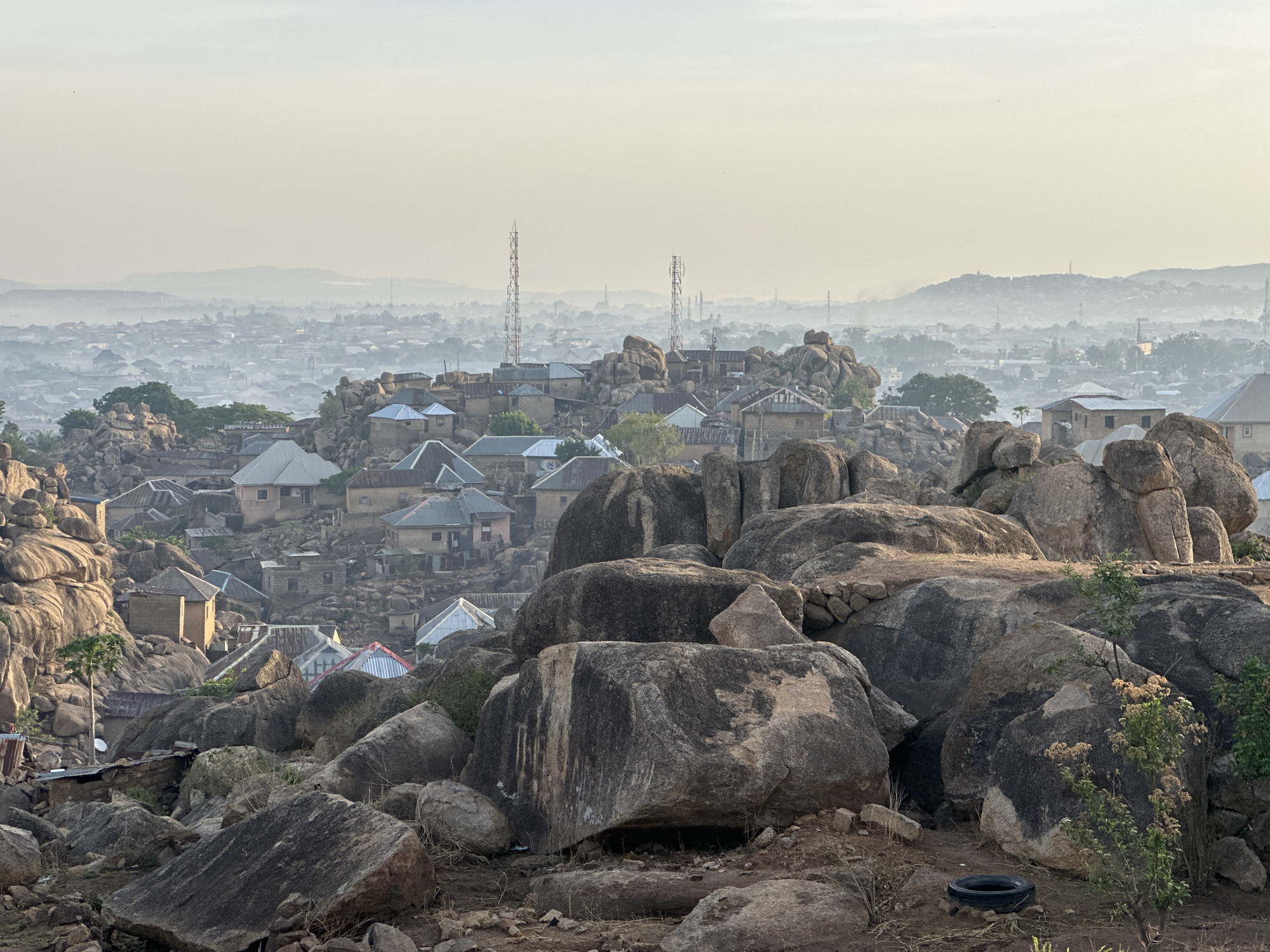 This panoramic image depicts a vast urban landscape in Jos, Nigeria, uniquely integrated with natural rock formations. The foreground is dominated by numerous large, weathered granite boulders of varying sizes, interspersed with dry earth and sparse, scrubby vegetation. In the bottom right corner, a single black tire lies on the ground.
In the midground, a residential area unfolds, where houses are built directly among and atop the massive rock formations. These homes feature a mix of corrugated metal and flat roofs, blending into the rugged terrain. A few trees, including a slender palm, dot the scene. Two prominent communication towers, likely cellular or radio masts, rise above the residential structures in the middle distance.
The background reveals a sprawling cityscape extending into the distance, blanketed by a soft, atmospheric haze. This haze lends a muted, almost ethereal quality to the distant buildings and the faint outlines of hills or mountains on the horizon. The diffused lighting suggests either an early morning or late afternoon setting. No people or specific activities are visible, presenting a serene, expansive view of the city. There is no visible text in the image.