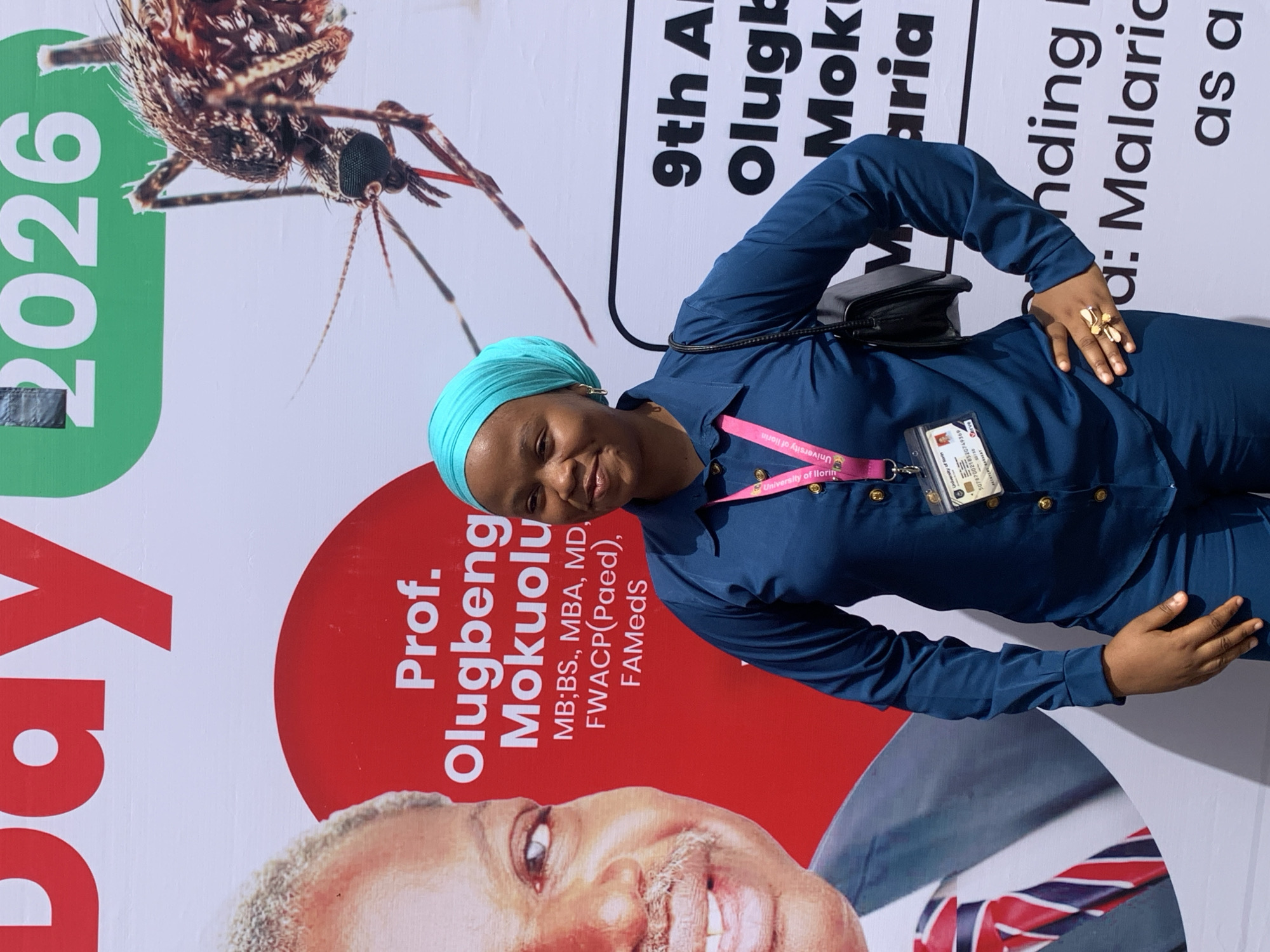 The image, rotated 90 degrees counter-clockwise, captures a smiling woman standing in front of a large event banner. She is dressed in a navy blue long-sleeved top and matching trousers, complemented by a light blue headwrap. A black shoulder bag is slung over her left shoulder, and a gold-colored ring with a butterfly design adorns her right hand, which rests on her hip. An ID badge, displaying "University of Ilorin" on its pink lanyard and a long numeric code "5078700218520749369" along with "Cerve", is clipped to her top.
The banner behind her is predominantly white with red and green accents. It features a large, detailed close-up of a mosquito, and the partially visible numbers "2026" in green. Text on the banner includes "Prof. Olugbenga Mokuolu" along with his qualifications: "MB;BS., MBA, MD, FWACP(Paed), FAMedS". Other partial texts visible are "9th A", "Olugb", "Mok", "aria", "nding I", "a: Malaria", and "as a", suggesting an event related to malaria, possibly a conference or annual gathering in Ilorin, Nigeria. A partial image of a smiling man, likely Professor Olugbenga Mokuolu, is also visible on the lower part of the banner, wearing a striped tie. No specific time of day or weather cues are evident.