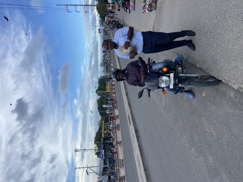 The image shows a scene in Dar es Salaam, Tanzania, taken from coordinates (latitude: -6.768, longitude: 39.226). Two men are standing on the side of a paved road, with a black motorcycle parked nearby. The man on the left is wearing a dark jacket, a helmet, and blue jeans. He is seated on the motorcycle, looking down. The man on the right is wearing a light blue shirt, dark pants, and black shoes. He is holding a brown paper bag and is looking at the camera. There are several buildings in the background, along with some traffic and street signs. The sky is blue with white clouds, and the weather appears to be sunny. There are multiple power lines in the sky and there are birds flying overhead. The location is likely a busy street in the city.