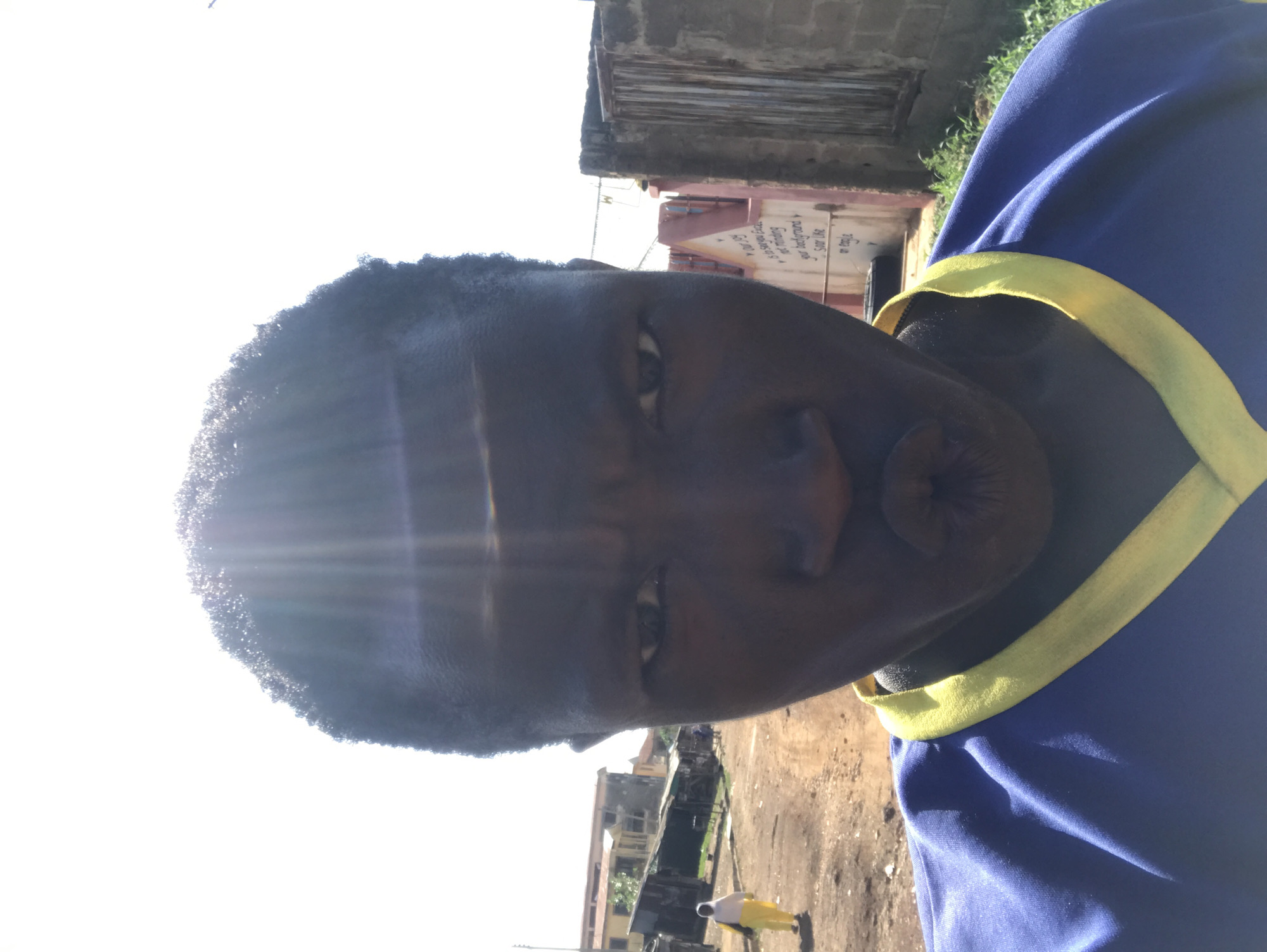 The image displays a close-up, rotated portrait of a person taking a selfie in an outdoor, brightly lit setting. The subject, who appears to be a young individual with dark skin and short, dark, curly hair, occupies the left side of the frame. They are wearing a blue shirt with a yellow trim. Their eyes are open and directed at the camera, and their lips are pursed in a pouting expression.
Strong sunlight is evident, creating significant lens flares and glare on the left side of the subject's head, indicating a clear, sunny day, likely around midday or early afternoon. The background reveals an urban or semi-urban environment with buildings and unpaved ground. To the right, behind the subject, a reddish-brown building with visible text inscribed vertically on its wall can be seen. Phrases discernible include "The Living God," "No meaning," "god background," and "SAVE LIFE."
The ground beneath the buildings appears to be dirt or gravel. In the lower right corner of the frame, a smaller figure of another person, dressed in yellow, is visible walking in the distance. The scene collectively suggests a location consistent with Mandate Estate, Ilorin, Nigeria.