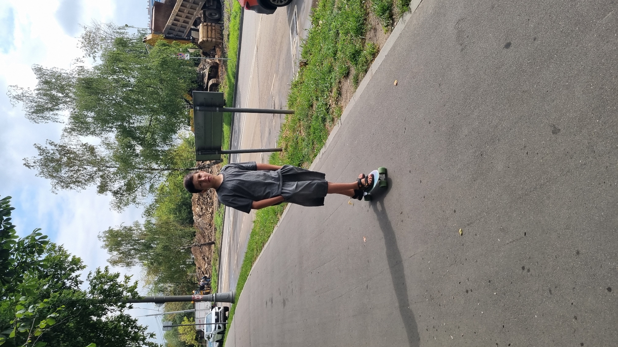 A boy stands on a skateboard on a paved pedestrian walkway next to a road. He is wearing a grey t-shirt and grey shorts, with his hands in his pockets. The skateboard is light green with black wheels. Behind the walkway, there is a grass verge and then a road with some construction activity. A large yellow dump truck is visible, along with piles of dirt and debris. Trees line the background, with a cloudy sky above. The scene appears to be in an urban area, possibly Moscow, Russia, given the presence of Russian signage on a post near the construction site.