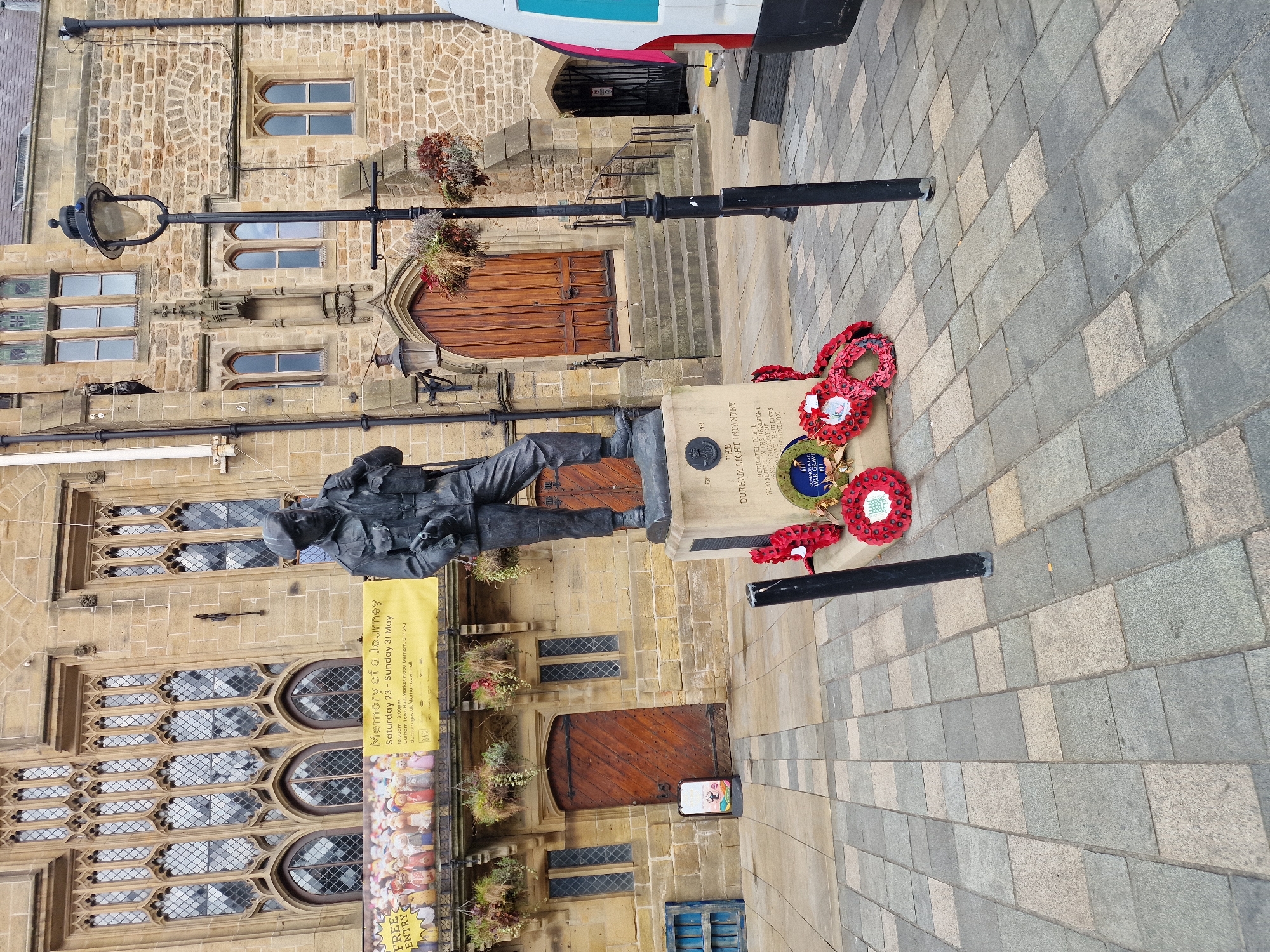 The image shows a war memorial in the city of Durham, United Kingdom. The statue of a soldier stands in front of Durham Town Hall. A banner to the left of the statue advertises an event called "Memory of a Journey" at the town hall from Saturday 23 to Sunday 31 May. Wreaths of poppies are placed on the memorial. The ground is paved with rectangular gray and tan stones. A lamp post is to the left of the memorial. A partial view of a vehicle is visible at the top right corner of the image.