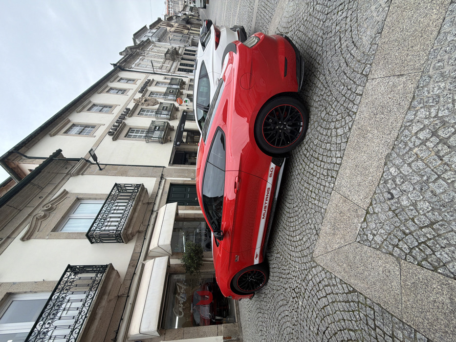 The image depicts a red Ford Mustang 5.0 coupe parked on a cobblestone street in Vila Real, Portugal. The vehicle is angled slightly towards the right, occupying the left side of the street, and features black multi-spoke wheels with red trim and a white "MUSTANG 5.0" decal along its lower side. Behind the Mustang, a white SUV is parked, followed by additional vehicles further down the street. The street surface consists of traditional Portuguese cobblestone, laid in a fanning pattern, with sections of lighter, larger granite slabs in the foreground.
The left side of the street is lined with multi-story buildings of traditional Portuguese architecture. The building immediately to the left of the Mustang features light-colored facades, ornate stone window surrounds, and black wrought iron balconies. Its ground floor houses a commercial establishment with large glass windows and two extended beige awnings. Reflections in the windows indicate a business named "Central" and an interior display featuring another red vehicle. Adjacent to this, another building with similar architectural characteristics displays a sign for "CALDEIRAS" and includes a visible ATM on its facade. The sky overhead is overcast. No individuals are visible in the scene.