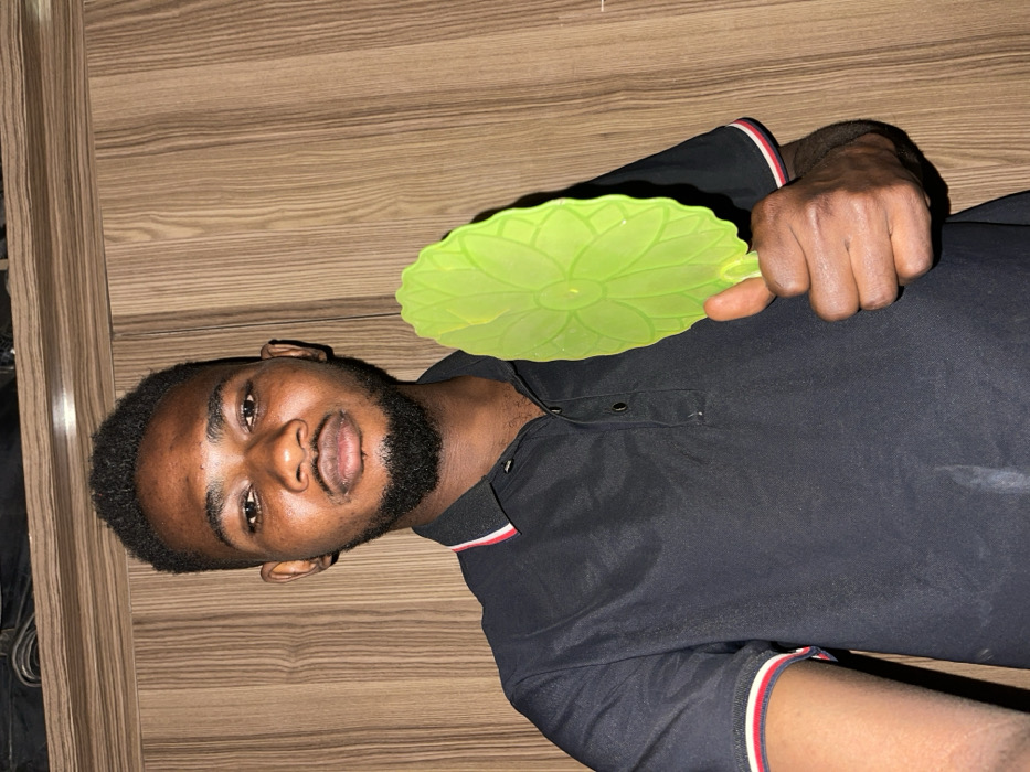A dark-skinned man with a beard poses in front of a wooden-looking background. He wears a black polo shirt with white and red accents on the sleeves and collar. He holds a green, lotus-shaped silicone lid in his right hand, displaying it towards the camera. The image is rotated, positioning the man on his side.