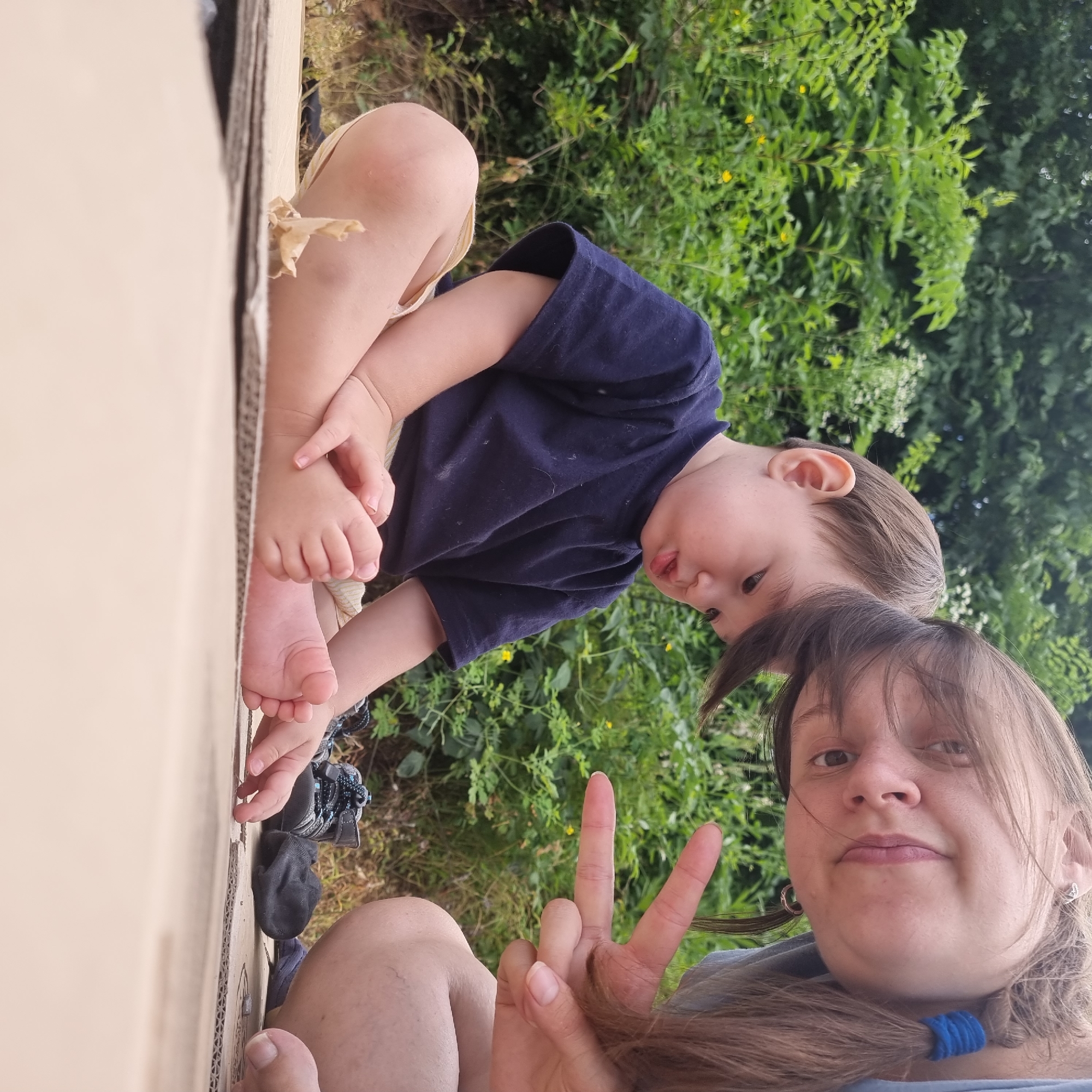 In this vertically oriented selfie, a woman and a young child are positioned within a large cardboard box. The woman is in the foreground, looking at the camera with a slight smile and holding up two fingers in a peace sign. Her hair is brown and falls across her face and shoulders, secured in a braid with a blue hair tie. She is wearing a light blue top.
Behind her, the child is leaning forward, resting on their knees and looking towards the woman. The child is wearing a navy blue t-shirt and light-colored shorts. Their small hands are visible, one resting on their knee and the other near their foot. The child's facial expression appears curious.
The setting is outdoors, with lush green foliage visible in the background, suggesting a natural environment. The lighting indicates it is daytime, likely with overcast or diffused light, as there are no harsh shadows. The subjects are within the confines of a large, brown cardboard box, which takes up a significant portion of the left side of the frame. Parts of the child's feet and legs are visible, as are the woman's legs and feet, suggesting they are both seated. A black sock and a grey and blue shoe are also visible on the ground near the child's feet. There is no visible text in the image. The overall mood appears relaxed and playful, as if the subjects are enjoying a moment of imaginative play.