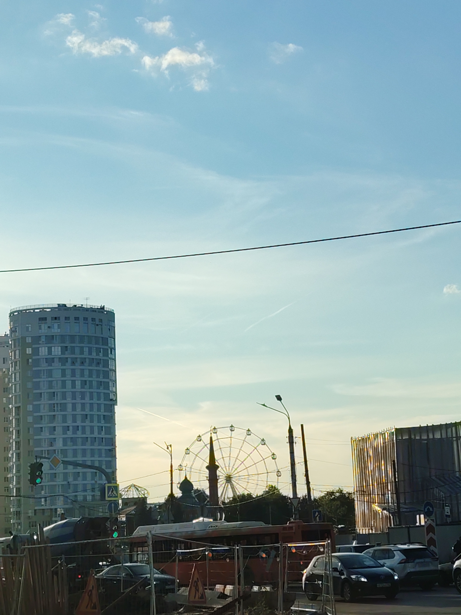 This is a full-frame, medium-shot, eye-level, daylight, exterior photograph taken from a street level perspective. In the background, a tall, cylindrical, modern building with many windows dominates the left side of the frame. To the right of this building, a Ferris wheel with radiating spokes is visible, with what appears to be a mosque-like structure in front of it, and another modern building with vertical, reflective panels to the far right. A street light with two arms is positioned between the Ferris wheel and the modern building. In the foreground, a street scene unfolds with several vehicles, including a concrete mixer truck, a red bus, and several cars. Construction barriers and orange traffic cones are present, suggesting ongoing road work. Traffic lights are visible, with at least one showing green. The sky is a pale blue with scattered, wispy clouds and contrails from aircraft. The scene depicts an urban environment in Nizhny Novgorod, Russia.