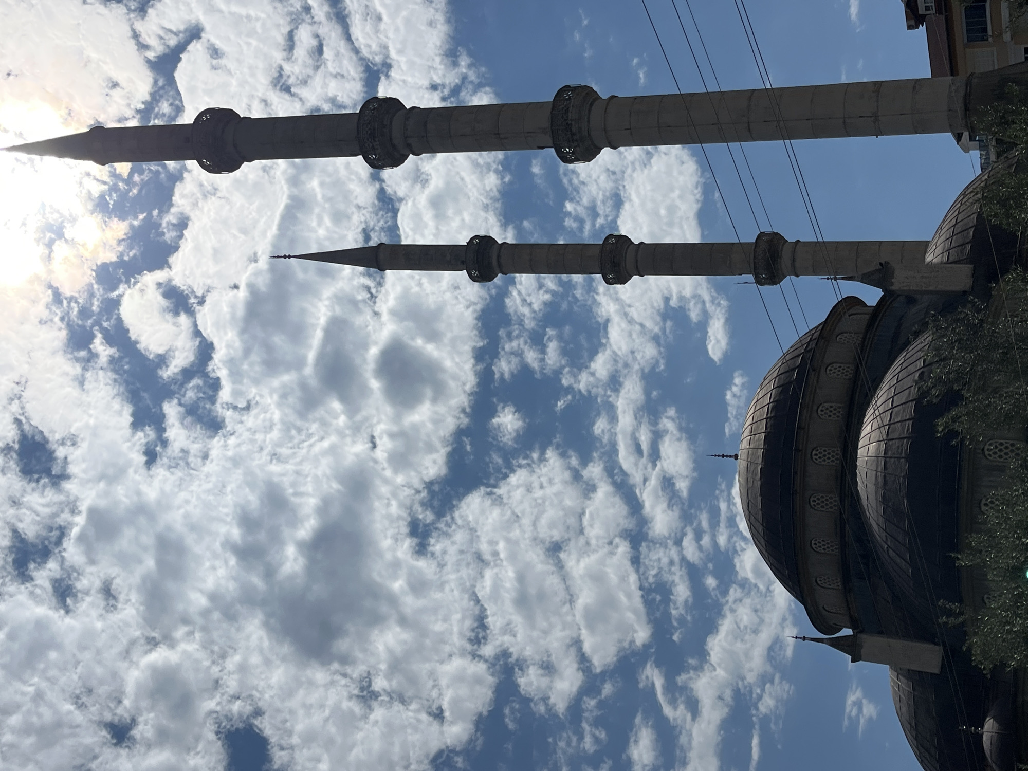 The image features a low-angle shot of a mosque in Alanya, Turkey, showcasing its architectural details against a backdrop of a blue sky with scattered white clouds. Two tall, cylindrical minarets with ornate ringed sections are prominently displayed, reaching towards the sky. The dome of the mosque, dark in color, is visible to the right. Power lines run across the sky between the minarets and the building. The building's location in the city of Alanya is supported by the style of architecture and the presence of other buildings visible in the background.