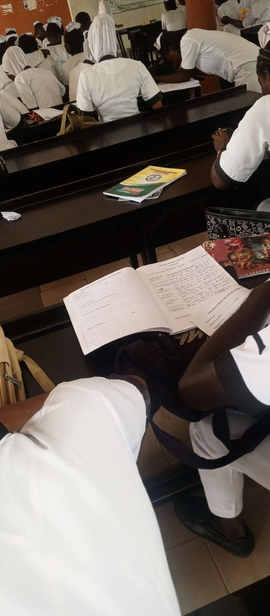 The image shows a group of young women in a classroom setting, likely in Kuson, Nigeria, based on the provided context. They are all wearing white uniforms, characteristic of nursing or medical students. Most of the women are seated at dark wooden desks, facing forward, with some appearing to be taking notes or listening to a lecture. One woman in the foreground is partially visible, wearing white pants and black shoes. Another woman is leaning over a desk, engaged in writing. Scattered on the desks are notebooks, pens, and other study materials, including a yellow booklet with green lettering. The overall atmosphere suggests an educational environment, possibly during a lesson or examination.