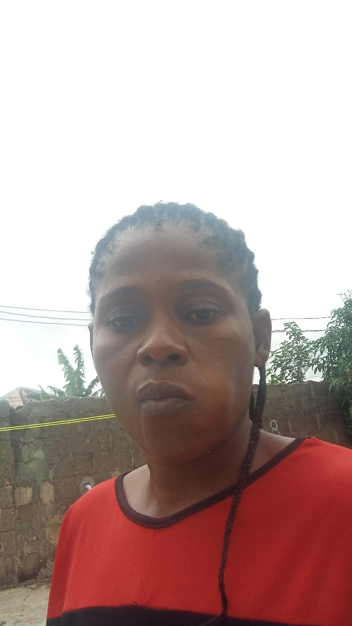 A woman with dark skin and braided hair is pictured in a close-up selfie. She is wearing a red and black t-shirt. She has dark eyes and lips that are pursed. Her cheeks are puffed out. In the background, there is a brick wall and some greenery. The sky is overcast. This image was taken in Rido, Nigeria.