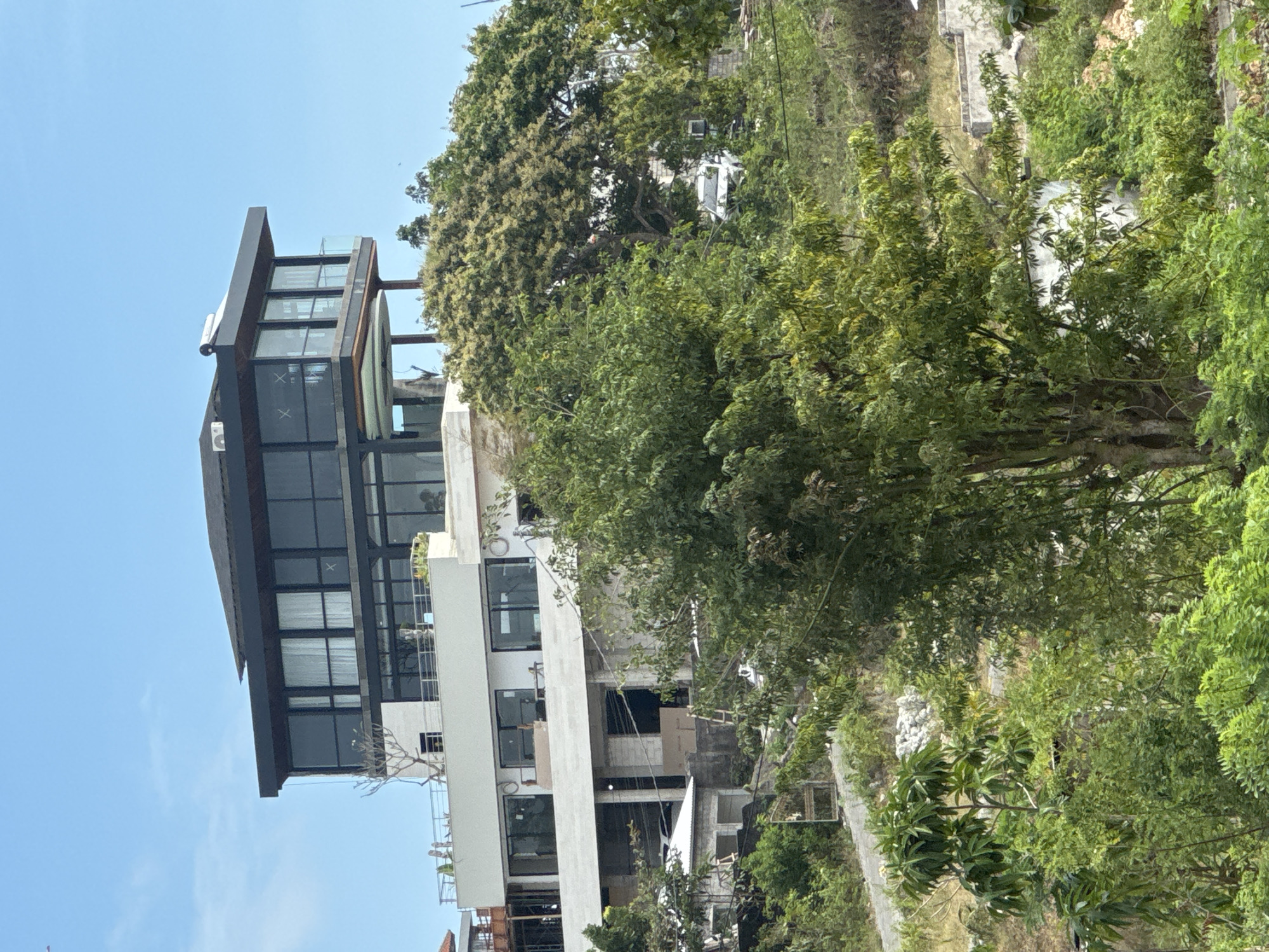 The image depicts a modern multi-story building situated on a hillside in Kuta Selatan, Indonesia. The building features extensive glass facades, particularly on the upper levels, which are cantilevered and topped with a dark, flat roof. Below the upper section, a lighter-colored facade with rectangular windows is visible, with an ongoing construction area on a lower level. The surrounding environment is lush with green foliage and trees, with a dirt road or path winding up the hill. In the background, glimpses of other structures and a white vehicle can be seen. The sky is clear and blue.