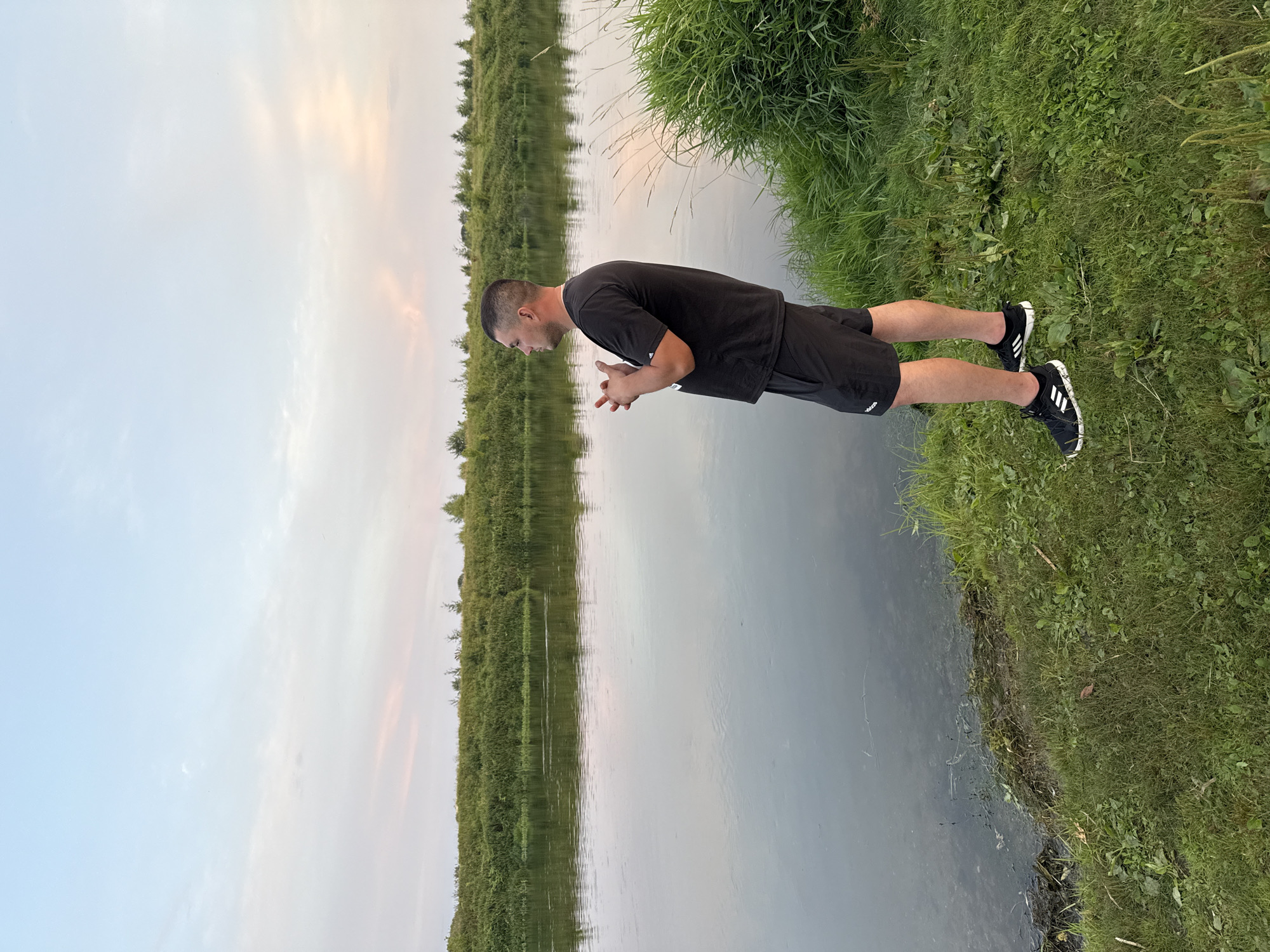 An adult male stands on the grassy bank of a wide, calm river, looking out over the water. He is wearing a black t-shirt and black shorts, and black athletic shoes. His hands are clasped in front of him. The river is surrounded by lush green vegetation on the opposite bank and tall grass on the immediate bank where the man is standing. The sky is a soft blue with hints of pink and orange from a sunset or sunrise. The scene appears to be in a natural, undeveloped area. The location is not definitively identifiable as Mahilyow, Belarus without further contextual information.