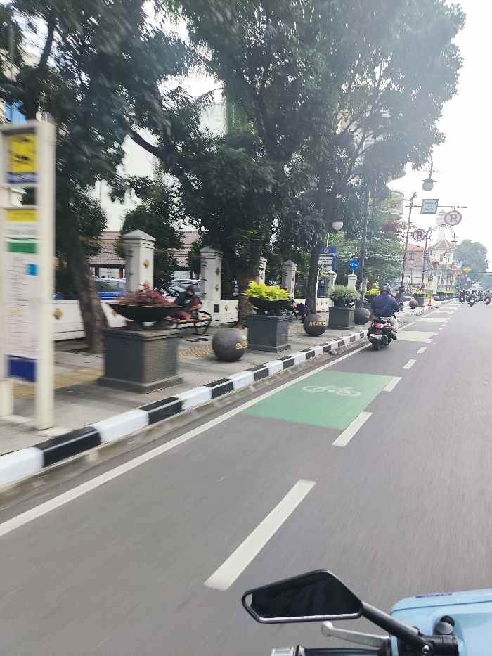 The image shows a street scene in Bandung, Indonesia, taken from a motorcycle traveling at a moderate speed. The coordinates indicate a location on Jalan Braga, a pedestrian-friendly street known for its cafes and restaurants. The road is paved with asphalt and has a bike lane marked with a green painted area on the right side of the frame. The motorcycle is traveling on the road, passing a sidewalk with a pedestrian walking on it. The sidewalk is lined with trees and benches. The scene is hazy, suggesting a cloudy day. The image is likely taken in the late afternoon or evening, judging from the angle of the sun and the shadows.