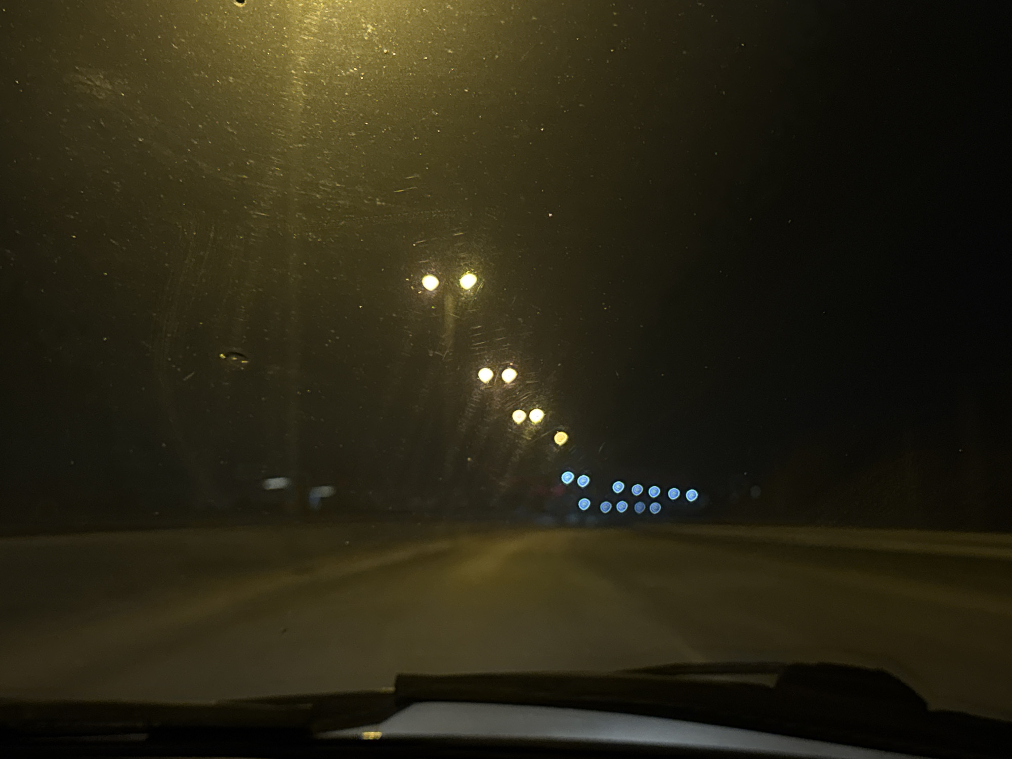 The image is a night-time view from inside a vehicle, presumably a car, driving on a road. Streetlights are visible along the side of the road, becoming more distant and blurred in the background. The windshield appears slightly dirty. The road is illuminated by the car's headlights and the streetlights. The wipers are visible at the bottom of the frame. The location is Кондратово, Russia, as stated.