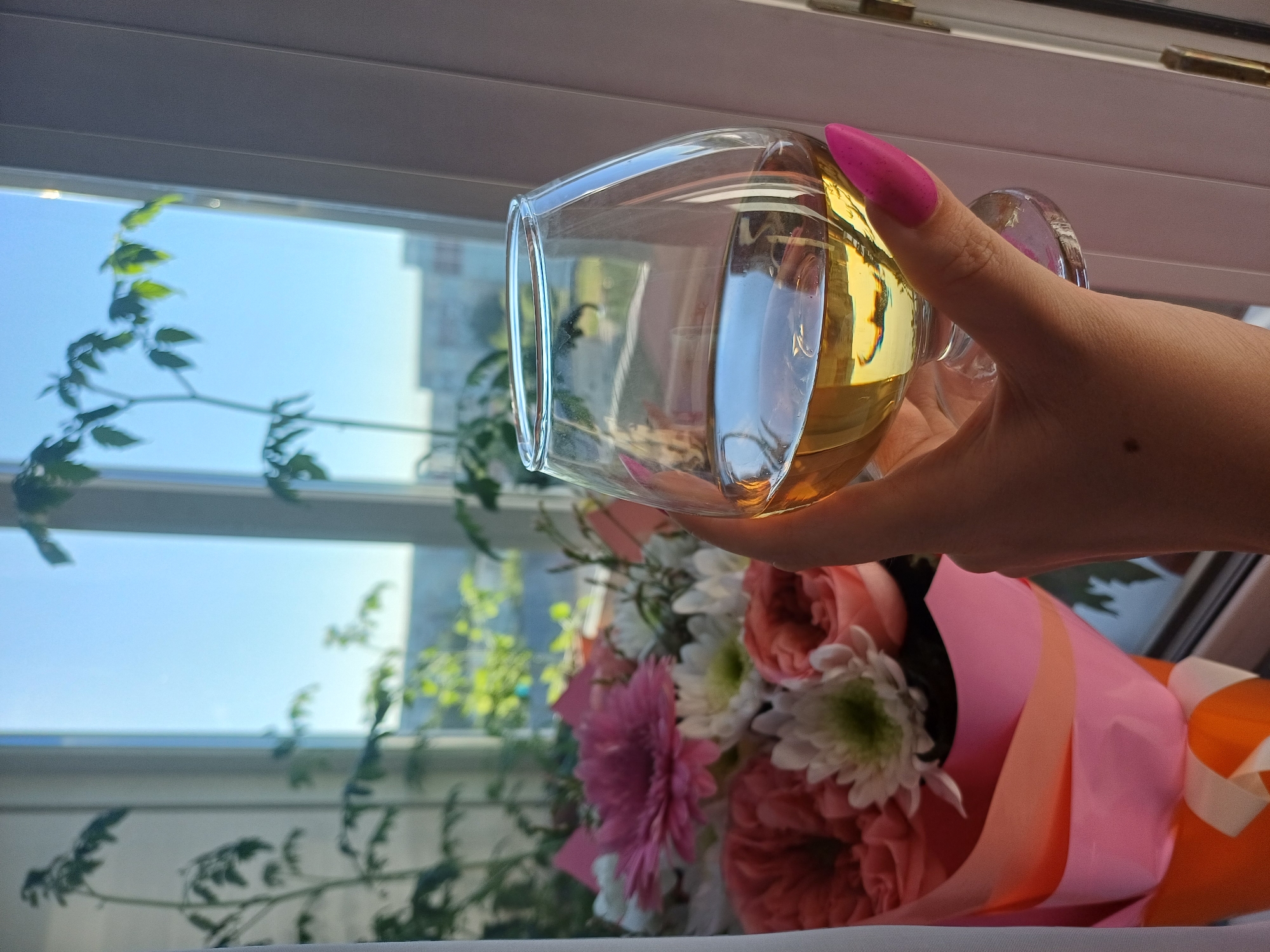 A close-up, rotated shot shows a hand holding a clear glass filled with a light-yellow liquid, likely wine or a similar beverage. The hand has bright pink, pointed fingernails. In the background, visible through a window with some plant vines, is a bouquet of flowers. The flowers are a mix of pink, white, and coral, with greenery. The bouquet is wrapped in pink and orange paper with ribbon accents. Sunlight streams in through the window, casting reflections on the glass and highlighting the flowers. The scene appears to be indoors, near a window, during the daytime. The overall mood is relaxed and possibly celebratory, with the wine and flowers suggesting a special occasion. No visible text is present in the image.