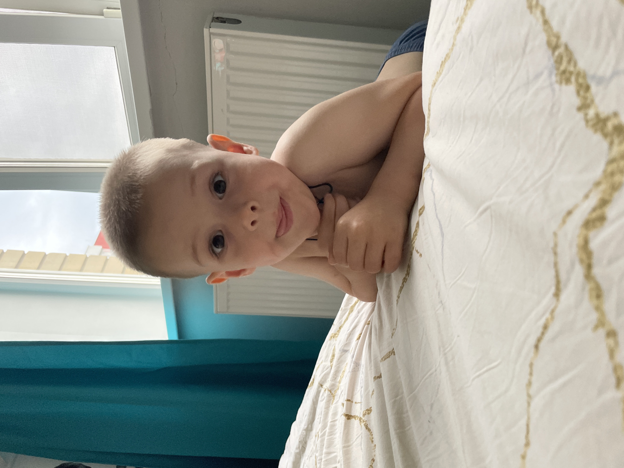 A young child is lying on a bed with white patterned bedding. The child is fair-skinned with short blonde hair, dark eyes, and a slightly open mouth. The child's arms are crossed, and they are looking directly at the camera. The background includes a window with white frames and a view of rooftops outside. There is a blue curtain to the left of the frame and a white radiator on the wall behind the child. The location is Unknown, Unknown.