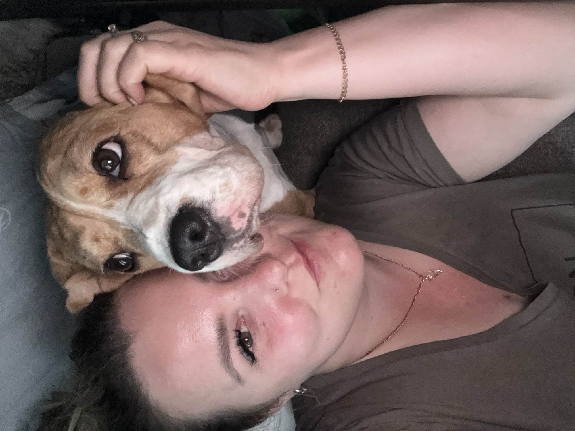 A selfie depicts a woman lying down with a dog resting its head on her forehead. The woman has fair skin, dark eyelashes, a nose piercing, and is wearing a tan shirt and gold necklace. She also has a ring on her left hand and a gold bracelet on her right wrist. The dog is light brown and white with large, expressive eyes and a black nose. The background is blurry and suggests an indoor setting, possibly a bed or couch. There is no information to suggest this image was taken in Марковский, Russia.