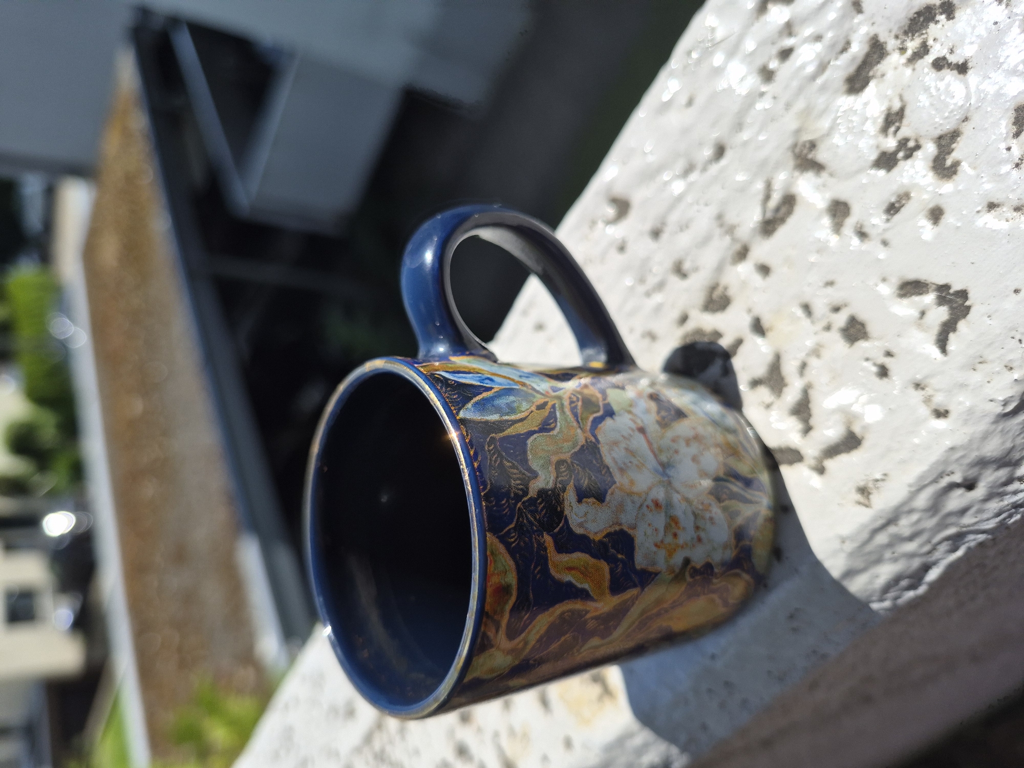 A close-up, eye-level shot features a blue mug with a floral design sitting on a textured white surface, likely a windowsill or balcony railing. The mug's interior is a dark blue, and the exterior showcases a pattern of gold, green, and white flowers against a navy background. The mug has a dark blue handle. The background is out of focus, showing a blurry cityscape with buildings and greenery. Sunlight casts a shadow of the mug on the white surface. The scene appears to be outdoors, possibly in a residential area of a city.