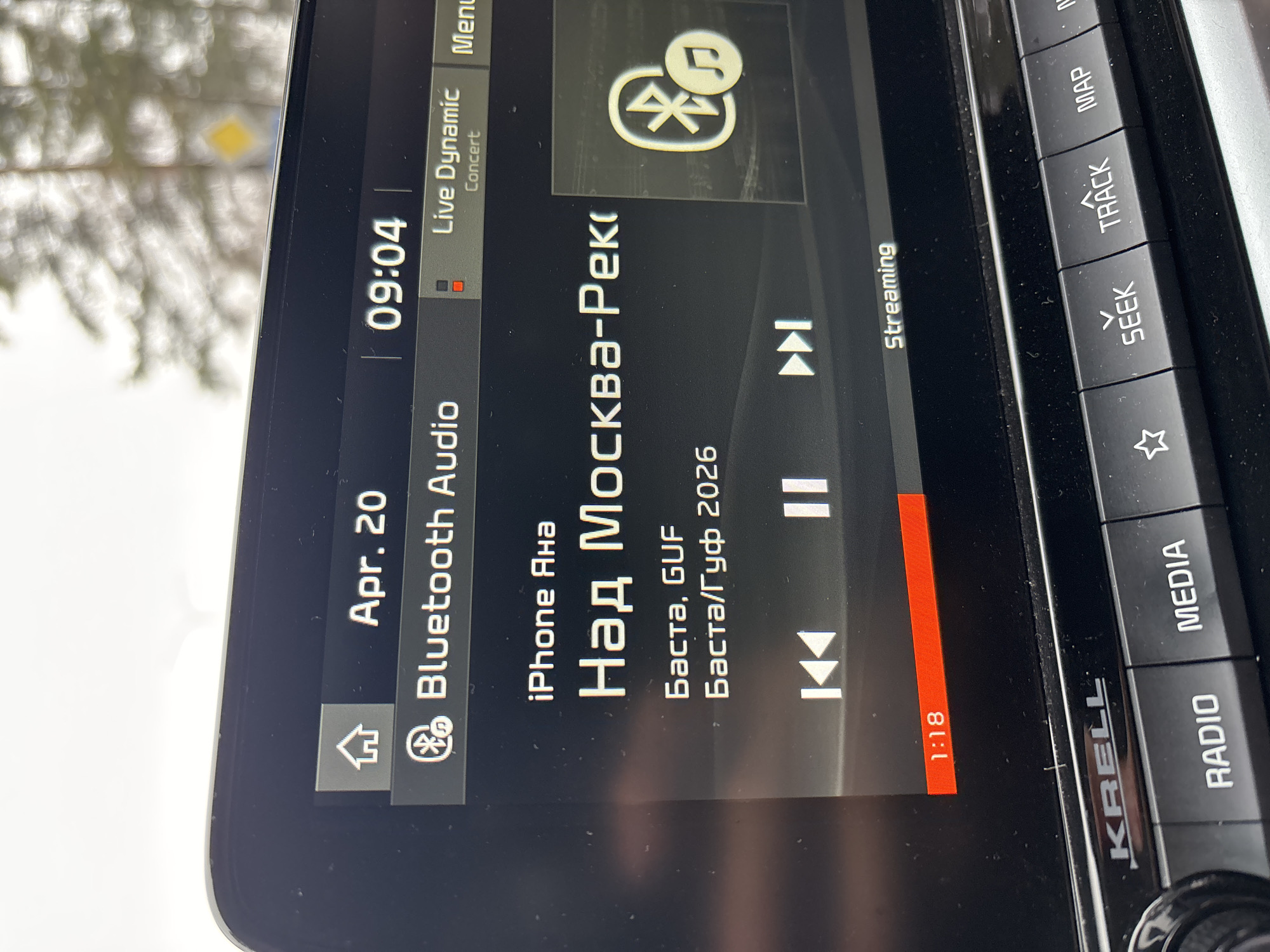The image shows a car's infotainment screen displaying music information. The date is April 20th and the time is 09:04. The audio source is Bluetooth, connected to an iPhone named "Яна". The song playing is "Над Москва-Реко" by Баста, GUF, from the year 2026. The audio system is KRELL. The music is streaming and the current time of the song is 1:18. The buttons on the side of the screen indicate the options to select radio, media, seek, track, and map. There is no way to determine the location of Chaikovsky, Russia based on the image.