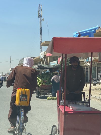 The scene depicts a street in Kandahar, Afghanistan, where life unfolds with its distinct characteristics. A man on a bicycle, wearing a brown robe and white head covering, carries a yellow canister strapped to the back of his bike. The frame's foreground captures another man wearing a head covering and operating a red food cart. Further along the street, a cart laden with watermelons is being tended to by another individual. In the background, a tall cell tower rises above buildings. The setting is arid and dusty, typical of the region.