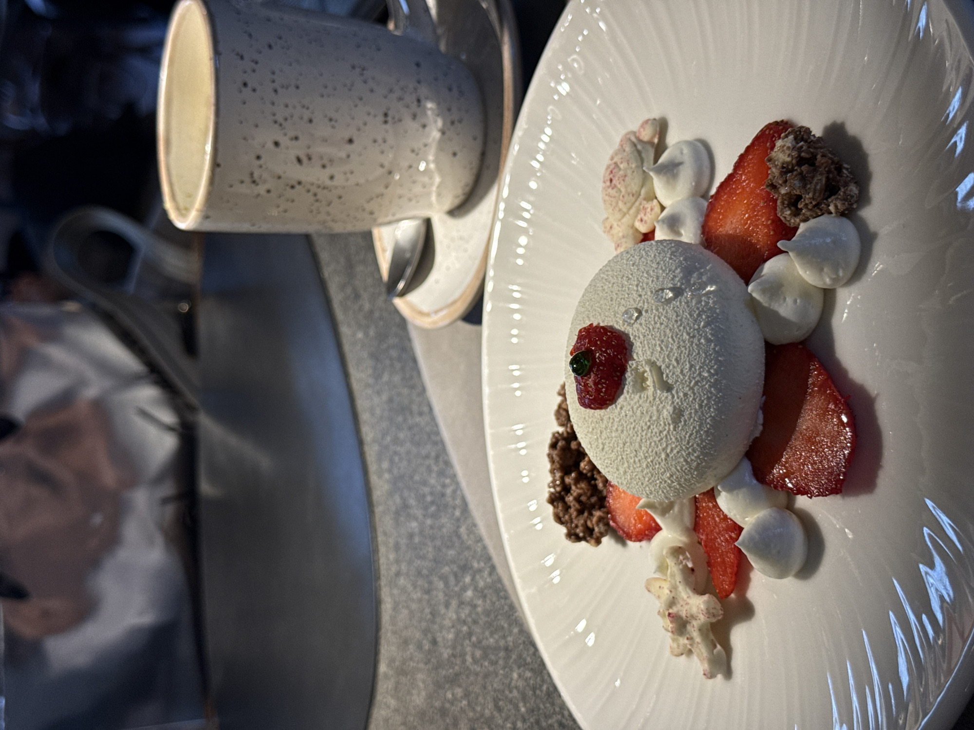 A close-up, angled shot shows a dessert plate and a mug, likely taken indoors. The dessert plate contains a white, dome-shaped sweet treat with a red decoration, garnished with sliced strawberries, dollops of cream, and what appear to be crumbled chocolate or nuts. A white mug with a speckled exterior sits next to the plate, partially obscuring the view. The setting appears to be a restaurant or cafe. There is no indication of the location being Krasnoyarsk, Russia. No people, specific actions or interactions are visible.