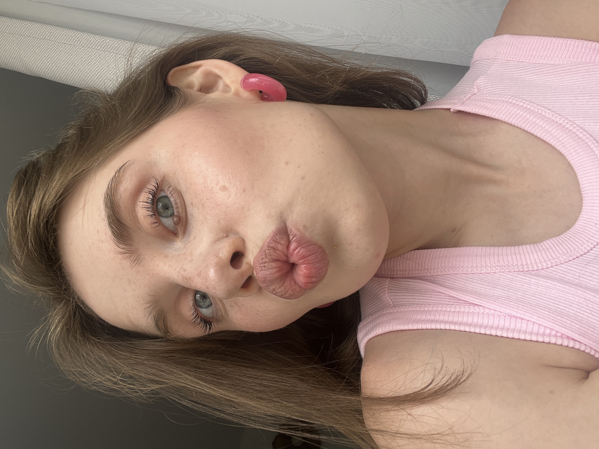 A young woman with light brown hair and freckles is captured in a close-up, head-and-shoulders shot. She is looking towards the camera with her lips pursed in a kiss pose. She has light blue eyes, styled eyebrows, and is wearing a pink tank top and pink hoop earrings. The background includes a plain gray wall and a white curtain. This image was taken in Chaikovsky, Russia.