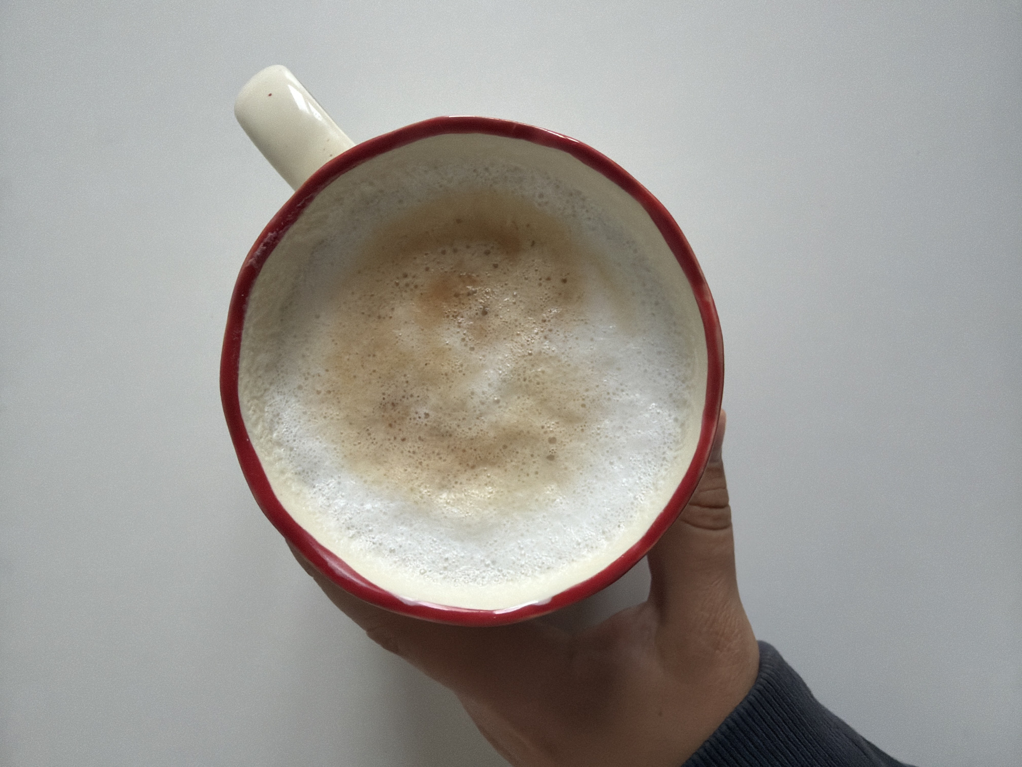 A top-down, medium shot shows a hand holding a red-rimmed white mug filled with foamy coffee. The coffee has a light brown crema. The mug has a white handle with a small red dot on it. The hand is holding the mug from the left side and is wearing a dark grey long-sleeved shirt. The background is a plain, light grey surface. The image appears to be taken indoors on a flat surface. The location is in the city of Фролы, Russia.