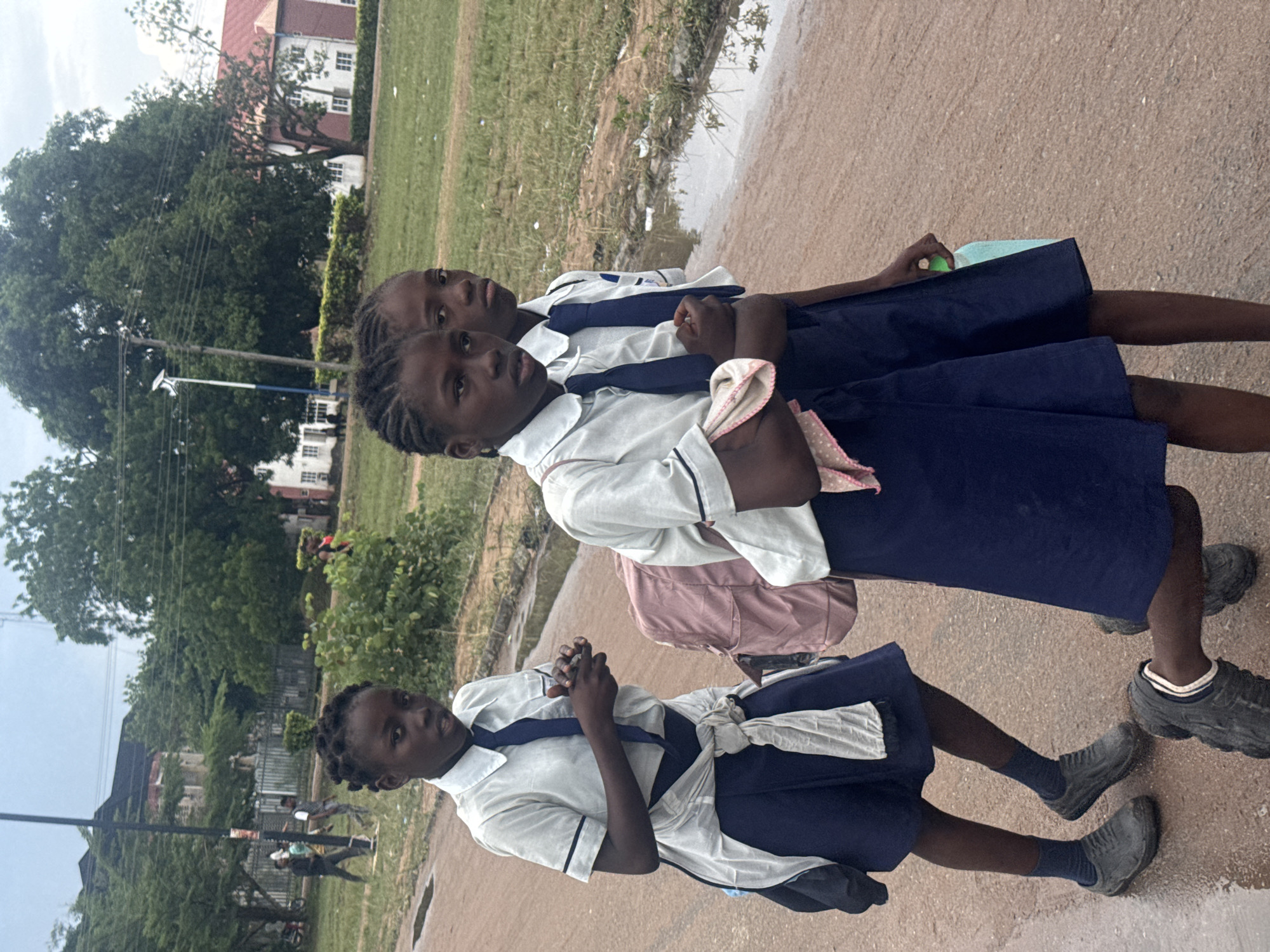 Three school-aged females, dressed in light-colored short-sleeved collared shirts with dark trim, dark ties, and dark blue skirts, walk on a reddish-brown unpaved road in the city of Ihiagwa, Nigeria. The road exhibits several puddles and wet patches, consistent with recent rainfall.
The individual on the left, positioned slightly forward, holds a small, dark rectangular object in her hands at chest level and has a light-colored garment tied around her waist. A dark bag is visible on her left shoulder. The central individual, partially behind the first, carries a pink backpack and has a light-colored, possibly polka-dotted, cloth draped over her right arm. The third individual, largely obscured by the central figure, holds a light green plastic bottle in her left hand. All three wear dark, closed-toe shoes that appear soiled, and have dark, braided hair.
The background consists of dense green foliage, large mature trees, and utility poles with overhead lines. Distant structures include a building with a dark roof on the left and a larger building with a distinct red roof and white walls on the right. Several adult males are visible walking in the mid-ground on the left side of the scene. The sky is overcast.