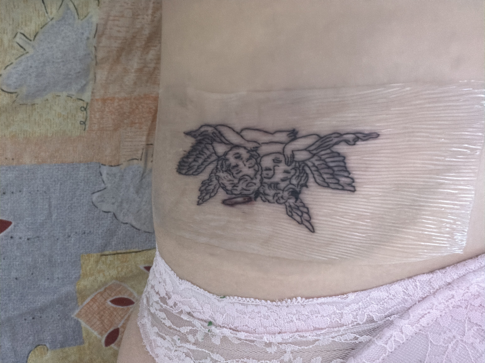 A close-up, indoor shot reveals the left side of a person's torso, featuring a freshly applied tattoo covered by a transparent protective film. The tattoo, rendered in black ink, depicts two winged cherubs or putti lying intertwined, facing each other. One cherub is positioned slightly above and behind the other, both adorned with curly hair and classical drapery, detailed with fine lines. The tattoo is situated on the side of the person's body, likely the rib cage or upper hip area.
The transparent film covering the tattoo is somewhat crinkled and slightly reflective, indicating it's a healing bandage like Saniderm. Below the tattooed area, the top edge of light pink, lace-trimmed underwear is visible, suggesting a delicate garment. To the left and in the background, a patterned fabric, possibly a bedsheet or blanket, displays abstract shapes in muted tones of beige, orange, and gray, providing a soft, out-of-focus backdrop. The overall scene suggests a personal and private setting, likely in Veliko Tarnovo, Bulgaria. No text is visible in the image.