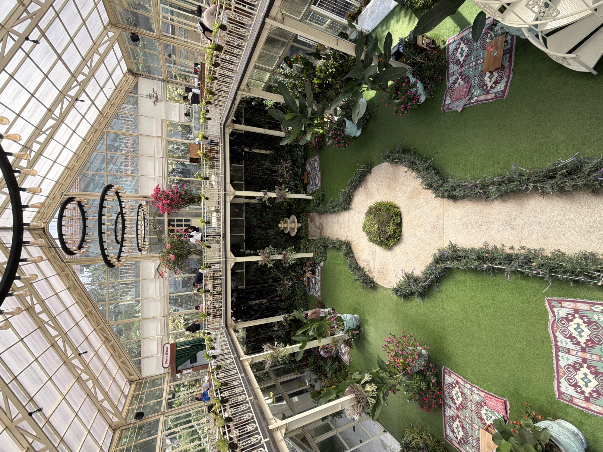 The image depicts the interior of a large, multi-level greenhouse cafe. The structure features a transparent glass roof and walls, allowing ample natural light to illuminate the space. The interior is designed with a central courtyard area covered in artificial turf.
On the ground level, there are several seating areas arranged on rugs, with plants and potted flowers surrounding them. A small fountain is visible in the center of the courtyard. A pathway made of light-colored stone winds through the artificial turf.
The upper levels consist of walkways and balconies overlooking the courtyard. These areas are lined with railings and also feature numerous potted plants. People are seen sitting at tables and conversing on these upper levels. A "Photo Booth" sign is visible on one of the upper walkways.
The overall atmosphere is lush and green, with a clear emphasis on bringing the outdoors inside. The cafe appears to be a popular gathering spot, with visitors enjoying the unique environment. The location is identified as a cafe in Baturiti, Indonesia.