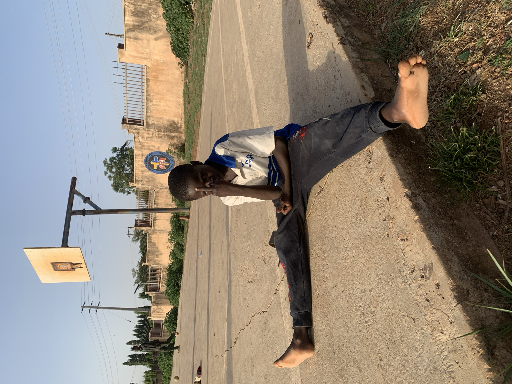 A young boy in a white and blue shirt and dark pants lies barefoot on a concrete surface next to a grassy edge. He is making a gesture with his hand, possibly a peace sign, while looking at the camera. In the background, a basketball hoop stands next to a building with a sign indicating a Christian fellowship. Utility poles and trees are also visible. The location is identified as Gura Top, Nigeria.