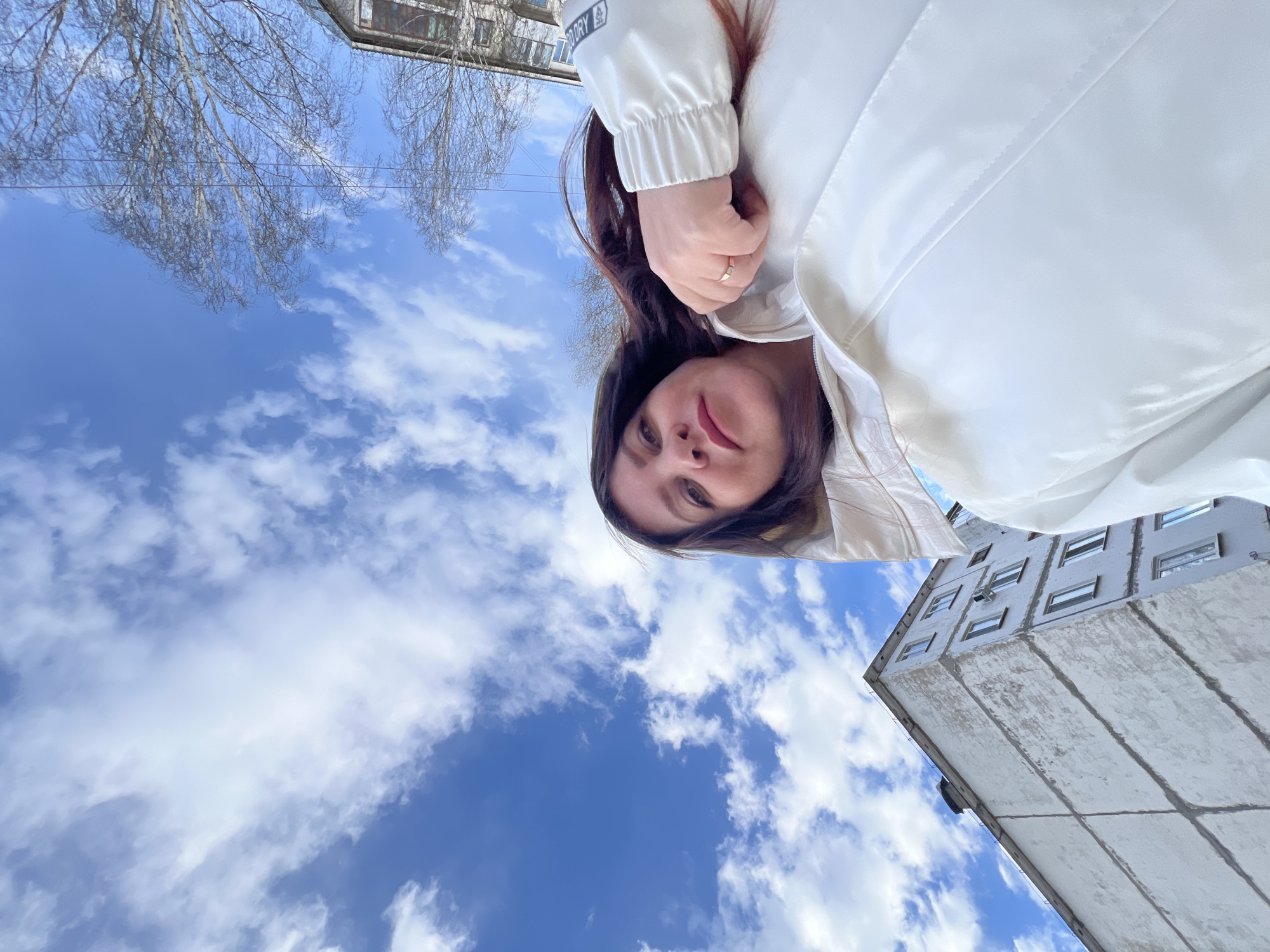 This is an outdoor, low-angle shot featuring a woman against a backdrop of a clear blue sky, white clouds, bare trees, and urban buildings, likely taken in Noviy, Russia. The image appears to be rotated approximately 90 degrees counter-clockwise from its natural orientation, suggesting an upward-looking perspective.
In the foreground, a woman with dark hair is visible from her head to her upper torso, looking directly towards the camera with a subtle, neutral to slightly content expression. She is wearing a clean white jacket, possibly a puffer or windbreaker style. A gold-toned ring with a small gem is visible on her right hand, which is lightly clasped. On her left sleeve, near the cuff, faint text can be seen, partially legible as "SPORT DRY," suggesting the jacket's material or brand.
The background is dominated by a vibrant blue sky dotted with fluffy white cumulus clouds, indicating bright, clear daylight. To the upper left, the intricate, leafless branches of a tree are silhouetted against the sky, suggesting either early spring or late autumn. On the right side of the frame, a tall, multi-story concrete building with numerous windows is visible, reflecting a utilitarian architectural style often found in post-Soviet urban landscapes. Another building with similar architecture is partially visible in the far background among the tree branches.
The overall mood is peaceful and bright, capturing a moment outdoors on a pleasant day. The low-angle perspective creates an expansive feel, emphasizing the sky and the surrounding environment.