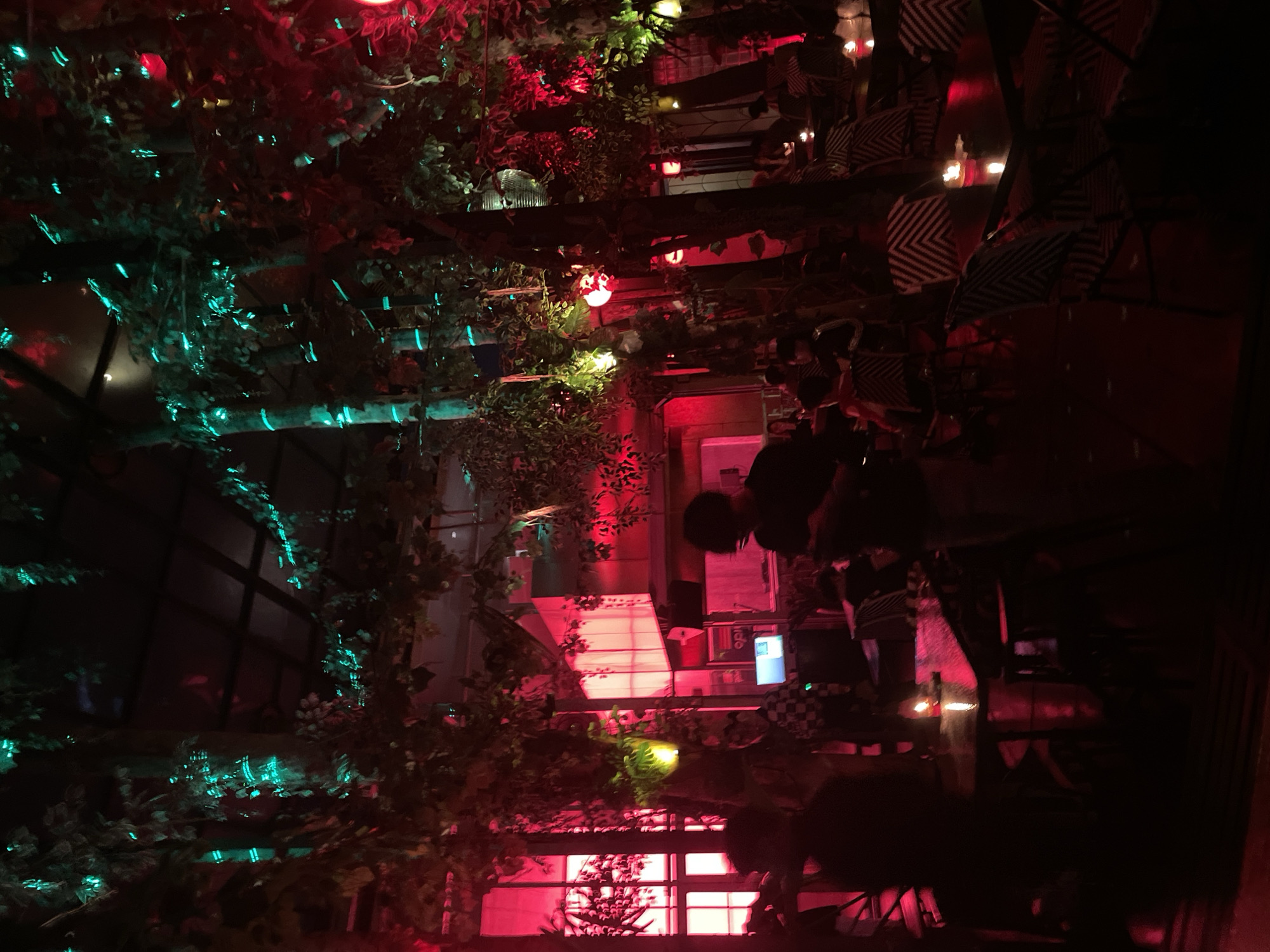 The image displays a dimly lit indoor or covered outdoor restaurant or lounge area, located in Bandung City, Indonesia. The scene is characterized by a prominent jungle or garden theme, featuring numerous trees and dense foliage throughout the space, illuminated by contrasting artificial lights.
Predominant lighting consists of red and teal-cyan hues. Intense red light emanates from large window-like structures on the left side of the frame and from several spherical red lanterns suspended at various heights. Teal-cyan light strips are wrapped around the trunks of multiple trees and integrated into the overhead foliage, creating a luminescent glow. Additional smaller green and yellow lights are interspersed within the greenery. Small, flickering candles provide warm, localized illumination on individual tables.
The environment includes a dark ceiling with visible structural elements and a large, rectangular, wall-mounted light panel emitting white light, situated on the mid-left. Numerous patrons are present, mostly seated at dark, rectangular tables equipped with small candles. Their figures are largely silhouetted against the ambient lighting. In the mid-ground, a person in dark attire stands, focused on a mobile device in their hands. To their left, another individual wearing a checkered shirt stands near a setup that includes a monitor displaying a blue interface, possibly associated with a DJ or sound system. The chairs feature a distinctive black and white chevron or zigzag pattern on their backs. Decorative red and yellow lanterns are suspended among the trees and foliage.