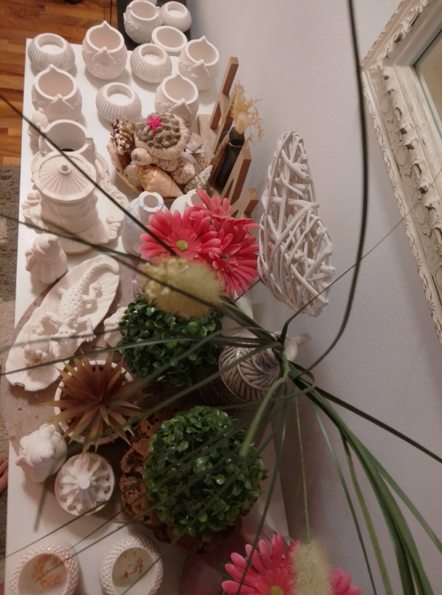 The image shows a collection of decorative objects, possibly on display or for sale, in a store in Wedel, Germany. The objects are primarily ceramic or natural materials and include a variety of white ceramic vessels, a ceramic lizard figurine on a leaf-shaped base, a cluster of seashells with a pink starfish, a rattan-like decorative sphere, and artificial flowers and plants. The items are arranged on a white surface, and the background includes a portion of a wall with a decorative ornate mirror frame. The lighting suggests an indoor setting. There are no people visible in the image.