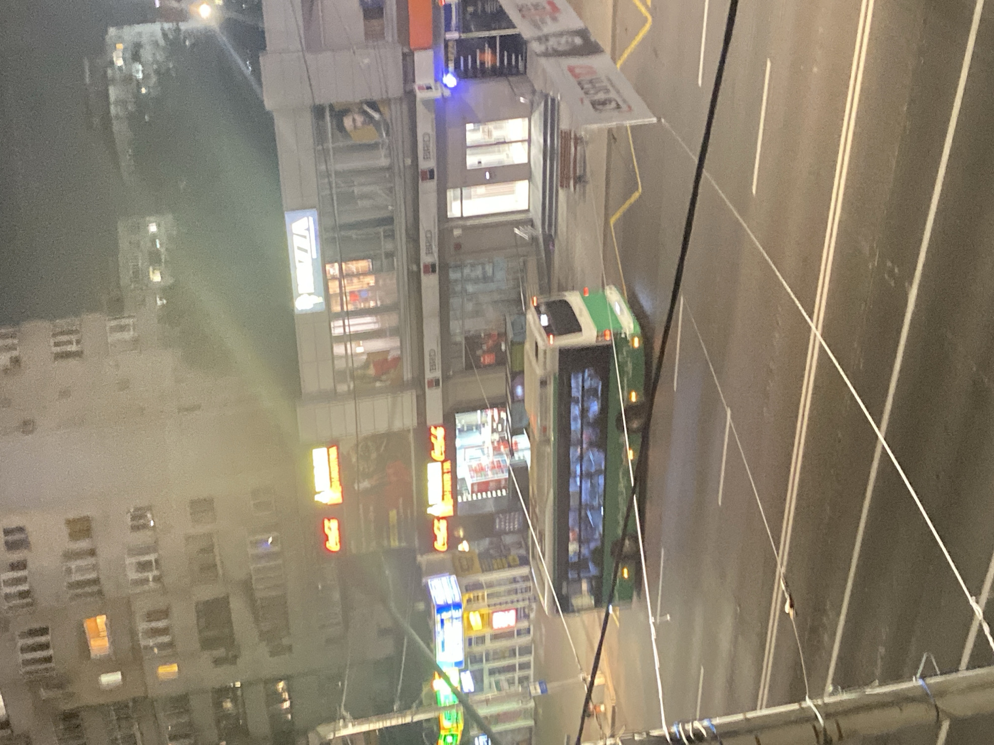 The image depicts a street scene at night in Iași, Romania. In the foreground, a trolleybus with a green and white exterior is traveling on the right side of the road. The bus has several windows, through which passengers can be seen. The road is asphalt with white lane markings. In the middle ground, a row of shops is visible. One shop has a sign that says "PIZZA" in blue and white letters. Another sign displays "Coca-Cola" in red and white lettering. The buildings have multiple floors and illuminated windows. In the background, taller buildings with many windows are visible, some of which are lit. The scene is illuminated by streetlights and the lights from the shops and buildings. Overhead wires, likely for the trolleybus, are strung across the scene.