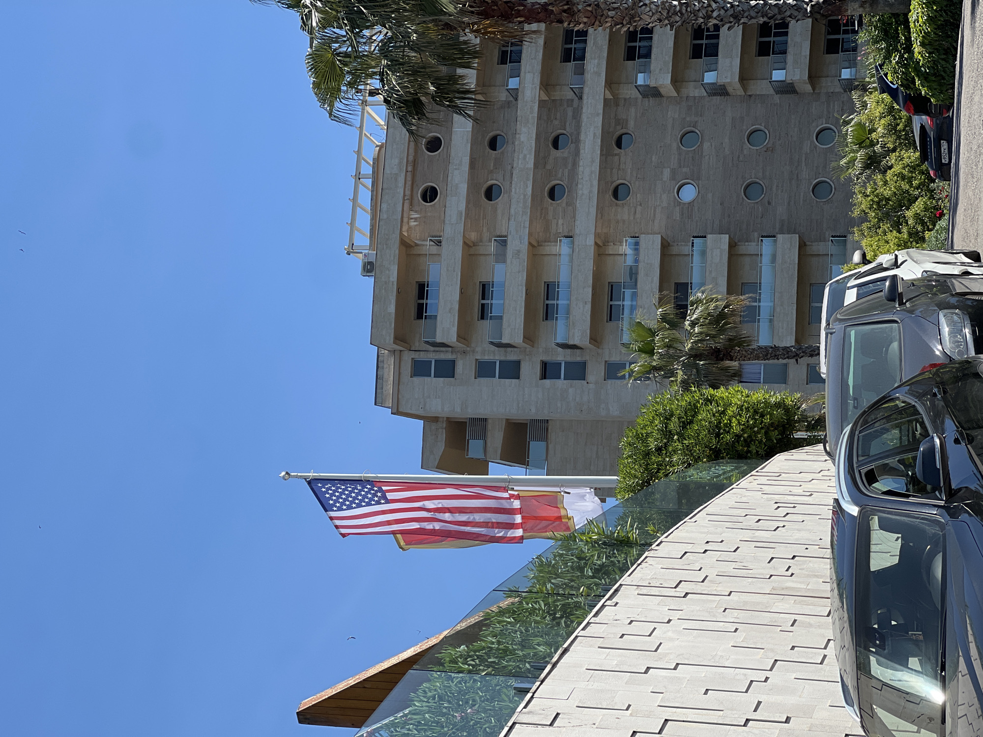 The image displays a sunny daytime scene featuring a modern building, flags, and parked cars, set against a clear blue sky.
The central subject is a multi-story building constructed from light-colored stone or concrete, exhibiting a contemporary architectural style. It features a repeating pattern of rectangular windows and distinct circular windows across various floors. Some levels are equipped with balconies that have glass railings. Palm trees flank the building, and dense green foliage is visible around its base and along a curved pathway.
In the foreground, an American flag waves prominently from a flagpole. Beneath it, another flag, predominantly red and white, is partially visible, consistent with the flag of Montenegro.
A light-colored, tiled pathway or ramp, edged with a glass barrier, curves across the lower right of the frame. Several vehicles are present; a dark SUV or van occupies the immediate foreground on the right, and other cars are parked or driving alongside the building. One dark car in the mid-ground has a partially visible license plate reading "H524 K*".
The sky is a vibrant, clear blue, indicating bright, sunny weather. A few small birds are visible in the expansive sky. The overall scene suggests a pleasant day in an urban or resort setting, consistent with the stated location of Budva, Montenegro.