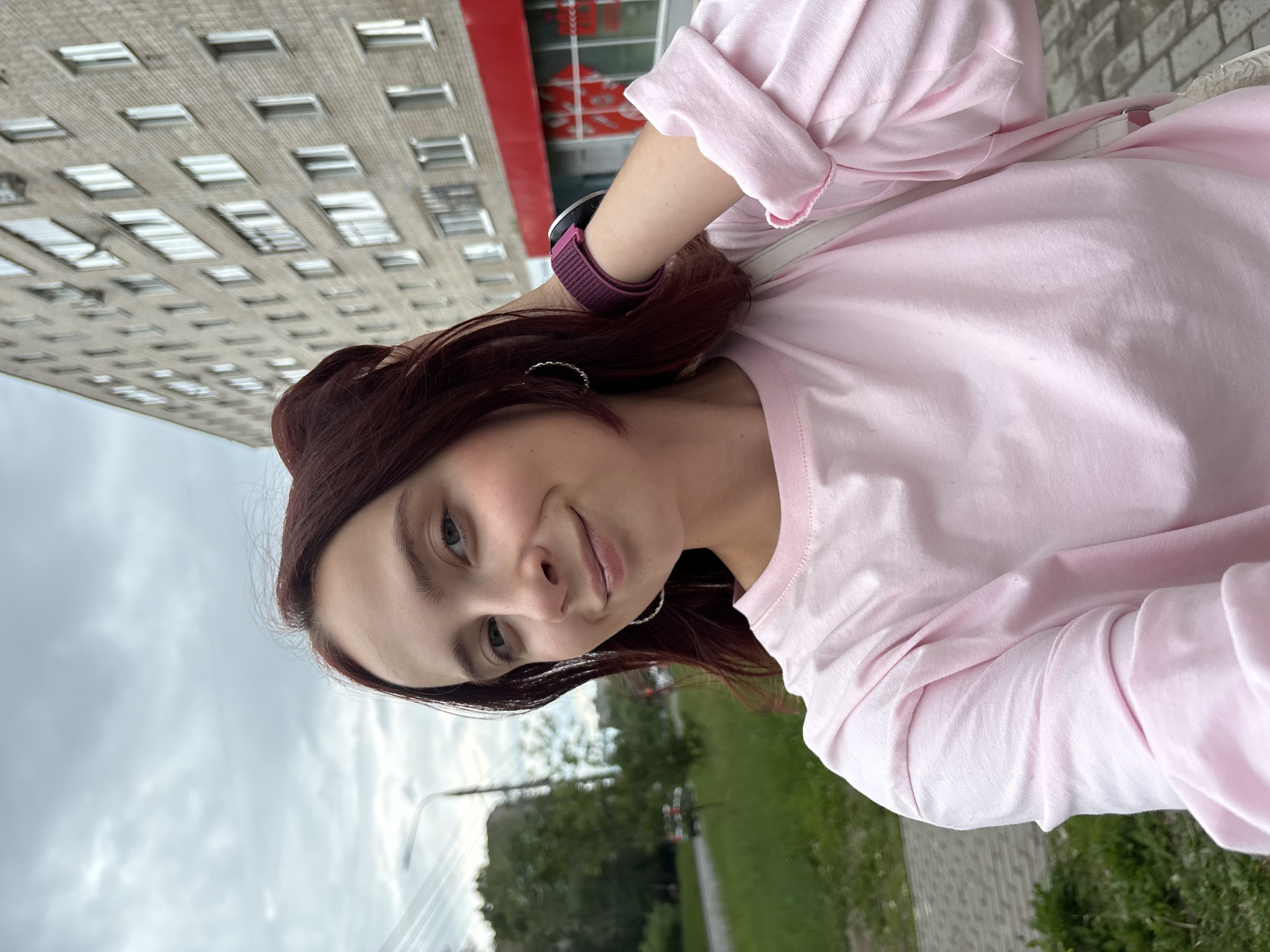 A young woman with auburn hair tied up in a bun and small hoop earrings is taking a selfie outdoors. She is wearing a light pink long-sleeved shirt and a dark purple smartwatch on her left wrist. In the background, a multi-story brick apartment building with many windows is visible, along with a street, some trees, and a cloudy sky. The scene appears to be on a sidewalk adjacent to a grassy area. The building and general urban setting suggest this photo was taken in the city of Izhevsk, Russia.