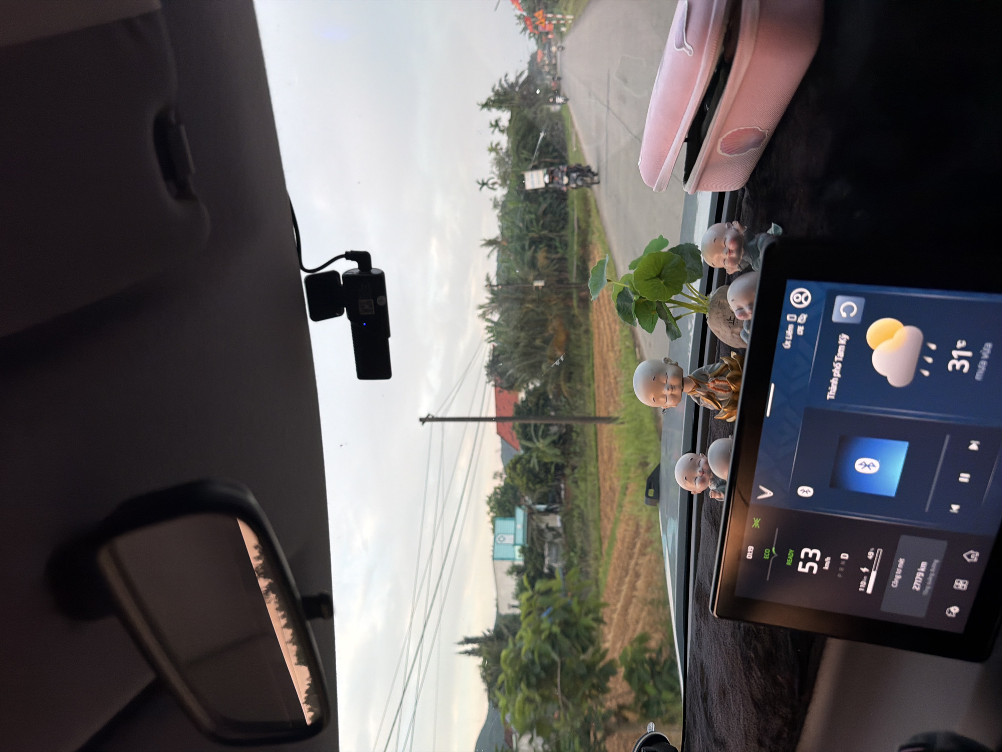 The image, rotated clockwise, provides an interior view from a car looking out onto a road in Bình Chương, Vietnam. On the left, a dashboard camera is mounted on the windshield, close to the rearview mirror. The dashboard is covered with a dark mat and adorned with three small monk figurines and a small potted plant. A pink pouch rests on the dashboard to the right.
Outside the car, a paved road extends into the distance, flanked by dense greenery, including palm trees, and what appears to be an agricultural field, possibly rice paddies. Power lines crisscross the sky, which is overcast, consistent with the weather information displayed. A motorcyclist is visible on the road ahead.
A car infotainment screen is prominent on the right, displaying digital information. The current time is "01:19". The car's status is "READY", with a speed of "53 km/h" and an odometer reading of "27179 km". The screen also shows the location as "Thành phố Tam Kỳ" (Tam Kỳ City), with a temperature of "31°C" and "mưa vừa" (light rain) indicated by a cloud and rain icon. A VinFast "V" logo is visible. Bluetooth is active, and a music player interface is shown, along with a battery status of "110-49%".