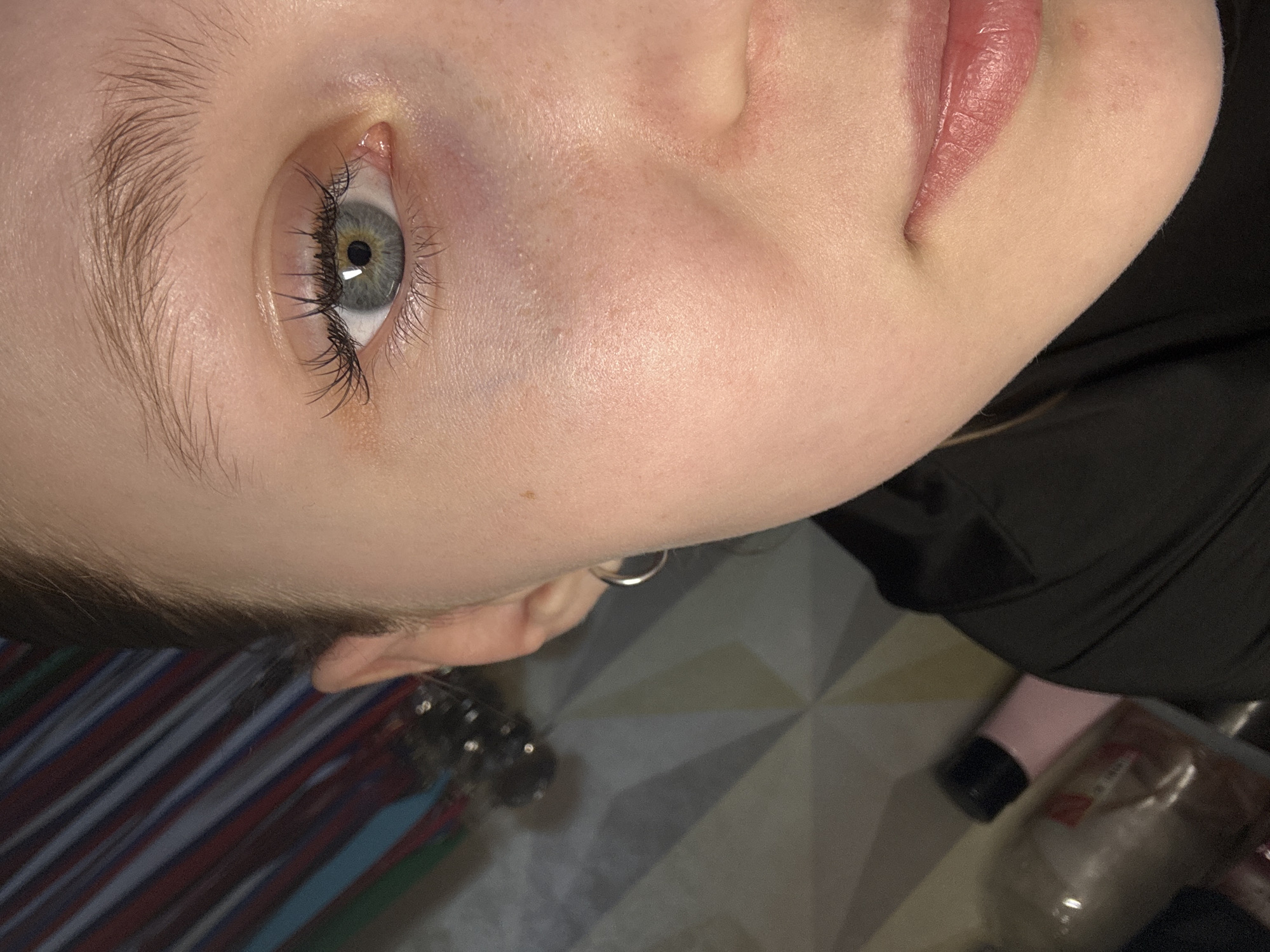 A close-up image shows a person's face, angled upward. The person has light skin, light eyebrows, and an eye with a light-colored iris and dark lashes. The person is wearing a small, silver hoop earring. Part of a black garment is visible in the upper right of the frame. In the background, there is a section of striped fabric and a patterned floor with some objects on it. The location and the city are not indicated in the image.