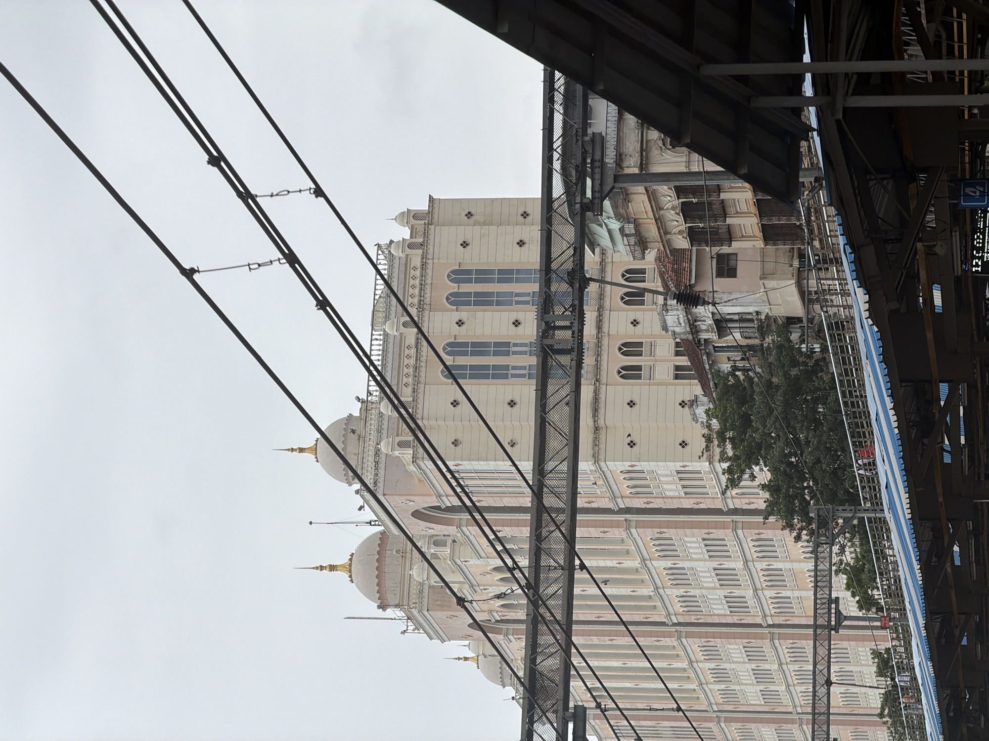The image captures a view of the Victoria Terminus, now Chhatrapati Shivaji Maharaj Terminus, in Mumbai, India. The foreground is dominated by overhead railway electrification wires, angled across the frame. Beneath these wires, a portion of a railway station roof, with a blue and white striped pattern, is visible, along with structural elements of the station. Behind the station structures, the ornate facade of the Victoria Terminus building rises, characterized by its Gothic Revival architecture. The building features a light pink and cream color palette, with multiple arched windows and decorative elements, including domes topped with golden spires. Several trees are interspersed between the station and the main building. The sky is overcast, suggesting a cloudy day. A few people are visible on a walkway or bridge in the midground, appearing as small figures against the backdrop of the architecture. The overall scene depicts a bustling urban environment with a focus on the historic railway station.