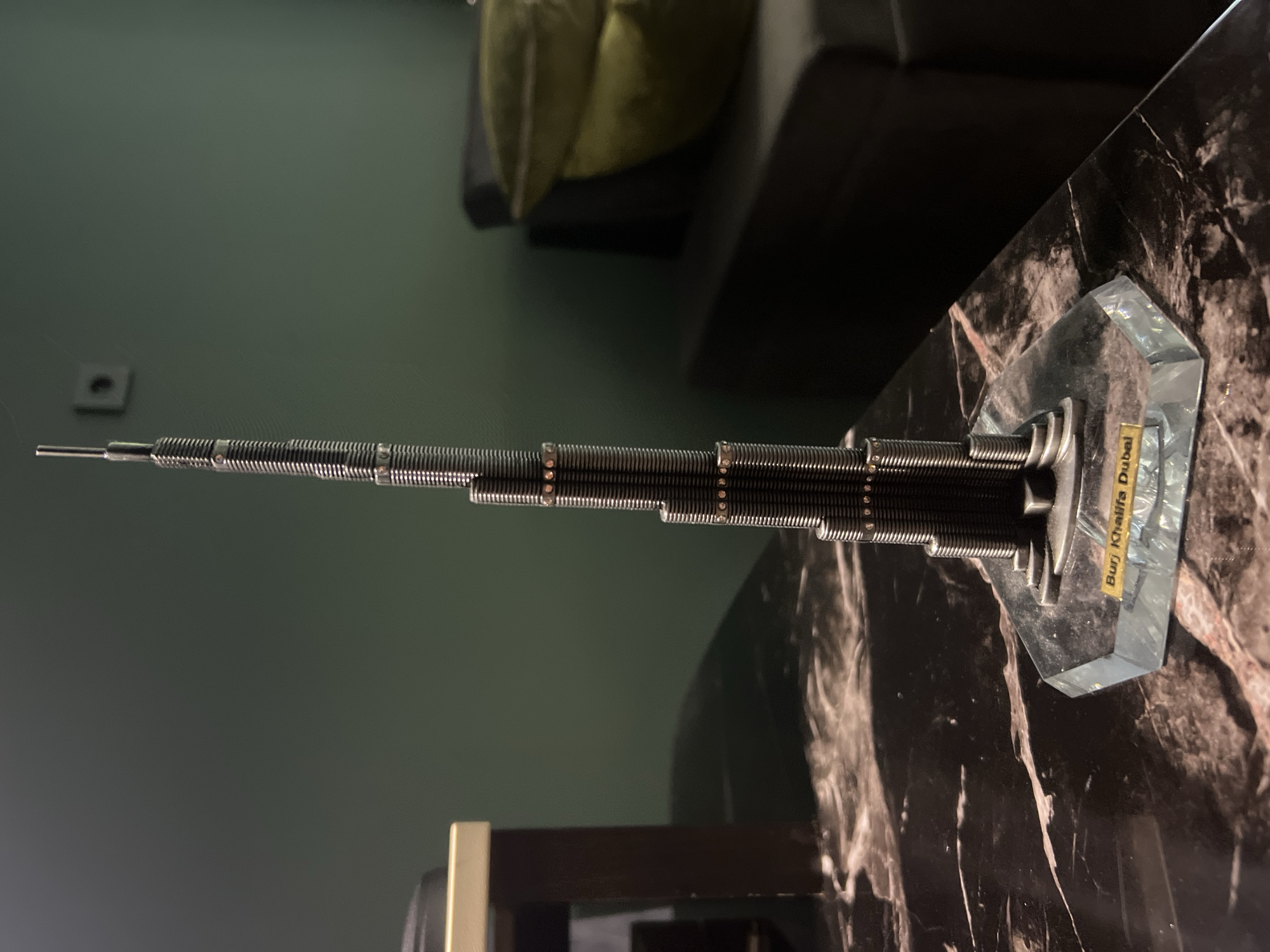 An eye-level medium shot shows a miniature model of the Burj Khalifa, a skyscraper, resting on a reflective, dark marble surface. The model is dark grey with horizontal ridged detailing and small, clear embellishments that appear to be crystals or rhinestones, encircling the tower at intervals. The base of the model is clear, faceted glass, with a small yellow plaque on its front face that reads "Burj Khalifa Dubai". To the left of the model, a portion of a dark wooden table leg and the edge of a light-colored surface are visible. In the background, a dark green wall is partially visible, with a small, dark square electrical outlet. To the right of the model, a dark-colored sofa with two dark green cushions is dimly lit. The lighting in the scene is dim, with the primary light source appearing to come from the front and slightly to the left, casting shadows.
