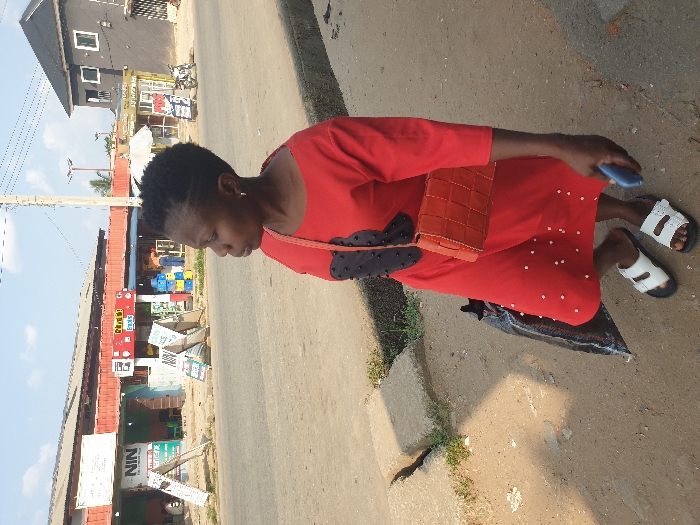 The image depicts a daytime street scene in Umuode, Nigeria. In the immediate foreground, a Black woman stands on a dirt and gravel roadside, facing right, slightly turned from the camera. She wears a red, mid-length dress with black accents on the chest and small white embellishments on the skirt, paired with white sandals with black soles. She carries an orange woven crossbody bag over her left shoulder and holds a blue smartphone in her right hand. A dark, possibly black, bag rests on the ground near her left leg.
Behind the woman, a paved asphalt road extends horizontally across the frame. On the far side of the road, to the left, a row of single-story commercial buildings with red corrugated metal roofs is visible. Prominent signage includes "NIN" and "GLO!", along with several A-frame signs. Yellow and blue plastic crates are stacked outside one establishment. To the right, further down the road, a dark-colored two-story building with white windows stands, followed by another commercial building with a white umbrella outside and a motorcycle parked nearby. The sky above is clear blue with minimal clouds. A utility pole and overhead wires are present in the background.