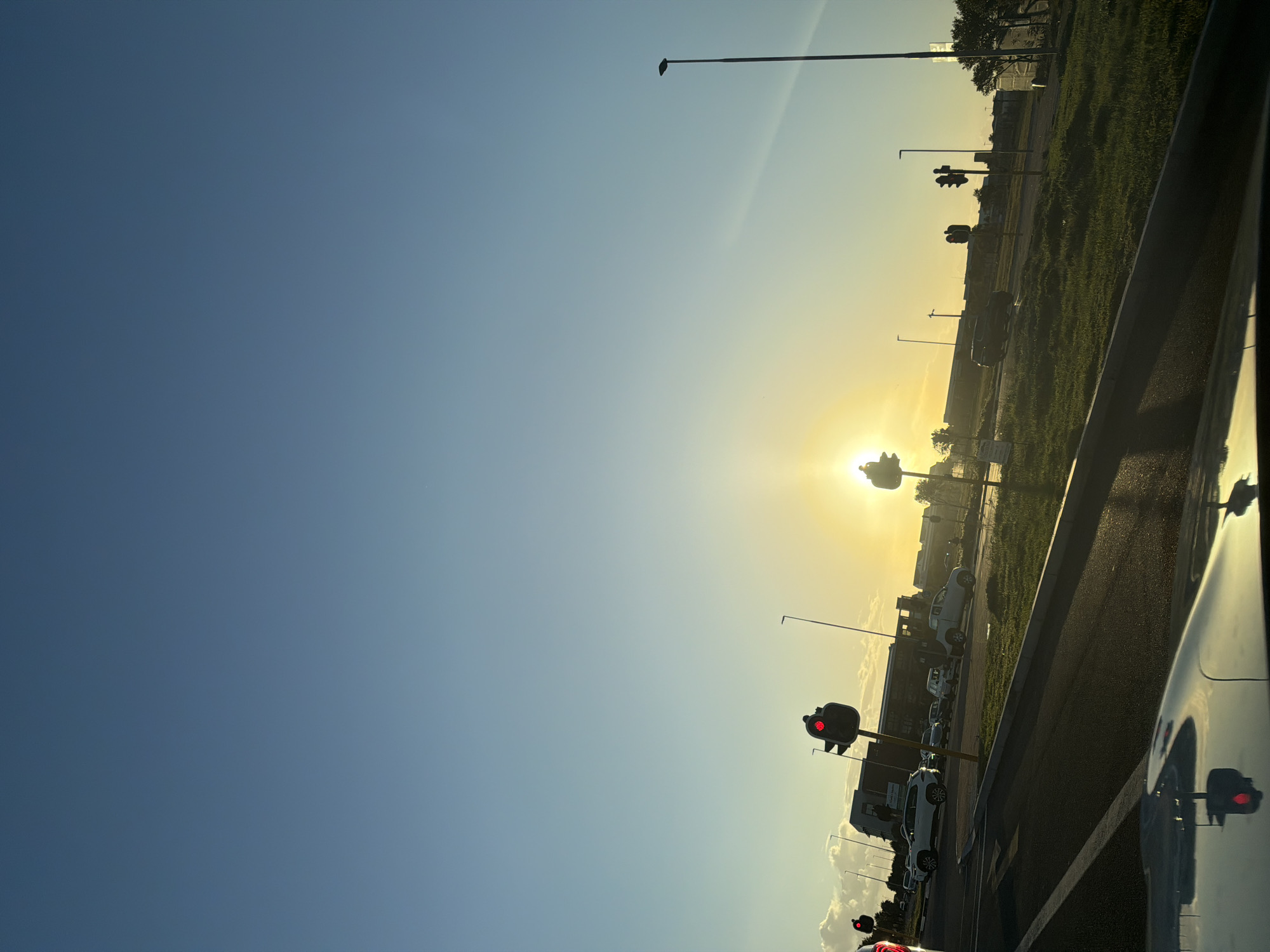 The image captures a street scene, likely taken from within a vehicle, during a sunrise or sunset, given the sun's position. The sky transitions from deep blue to a bright, golden-yellow hue near the horizon, where the sun shines intensely. The road stretches into the distance, bordered by a grassy area and buildings on one side. Several streetlights and traffic signals are visible along the roadside, their red lights indicating a stop. Cars are parked or lined up, suggesting an urban setting. The reflection of the traffic signal is captured by the car's window. I do not have enough information to determine the specific location in Cape Town, South Africa.