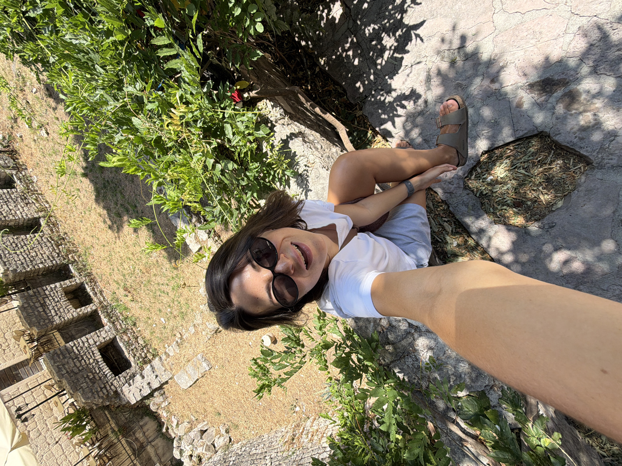 A young woman is taking a selfie from a high angle in an outdoor setting. She is wearing a white t-shirt, light blue shorts, and tan sandals. Her dark hair is styled down, and she has on large, dark sunglasses. She is smiling with her mouth open, and her right arm extends towards the camera, holding the phone.
The background shows a mix of stone structures and lush green foliage under a bright sun. The stone buildings appear old, with visible brickwork and arched openings. Dry, light-colored grass and debris cover the ground in some areas. The woman is seated on a stone surface, with shadows from trees dappling the ground around her. To the left of the frame, a portion of a large tree trunk is visible.
The scene appears to be within the historic Old Town of Budva, Montenegro, possibly in a courtyard or garden area adjacent to the ancient fortifications. The presence of stone architecture, Mediterranean vegetation, and the general ambiance suggest this location.